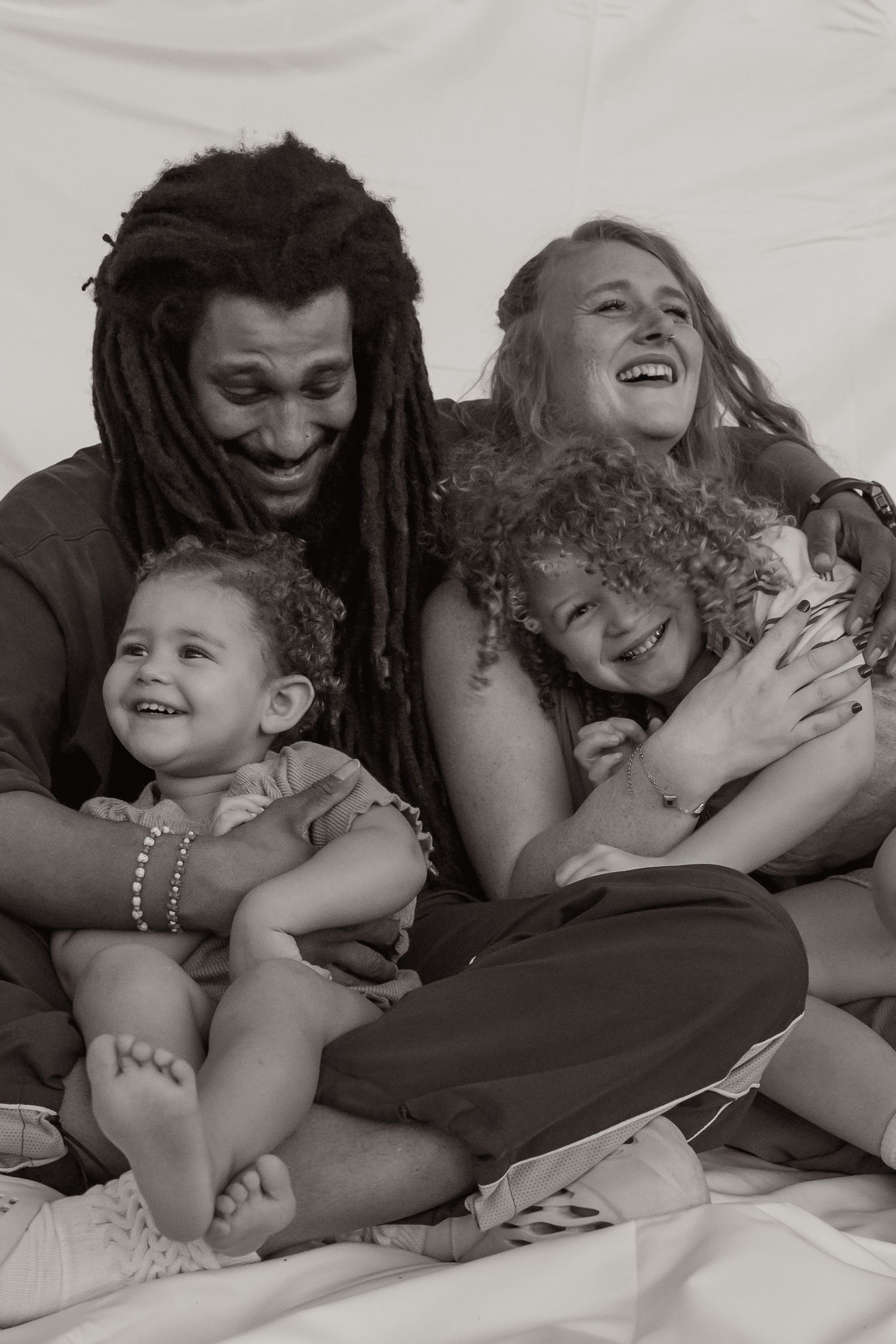 A joyful interracial family laughs together in a black and white portrait with two young curly-haired children. Taken by Row And Co in FXBG