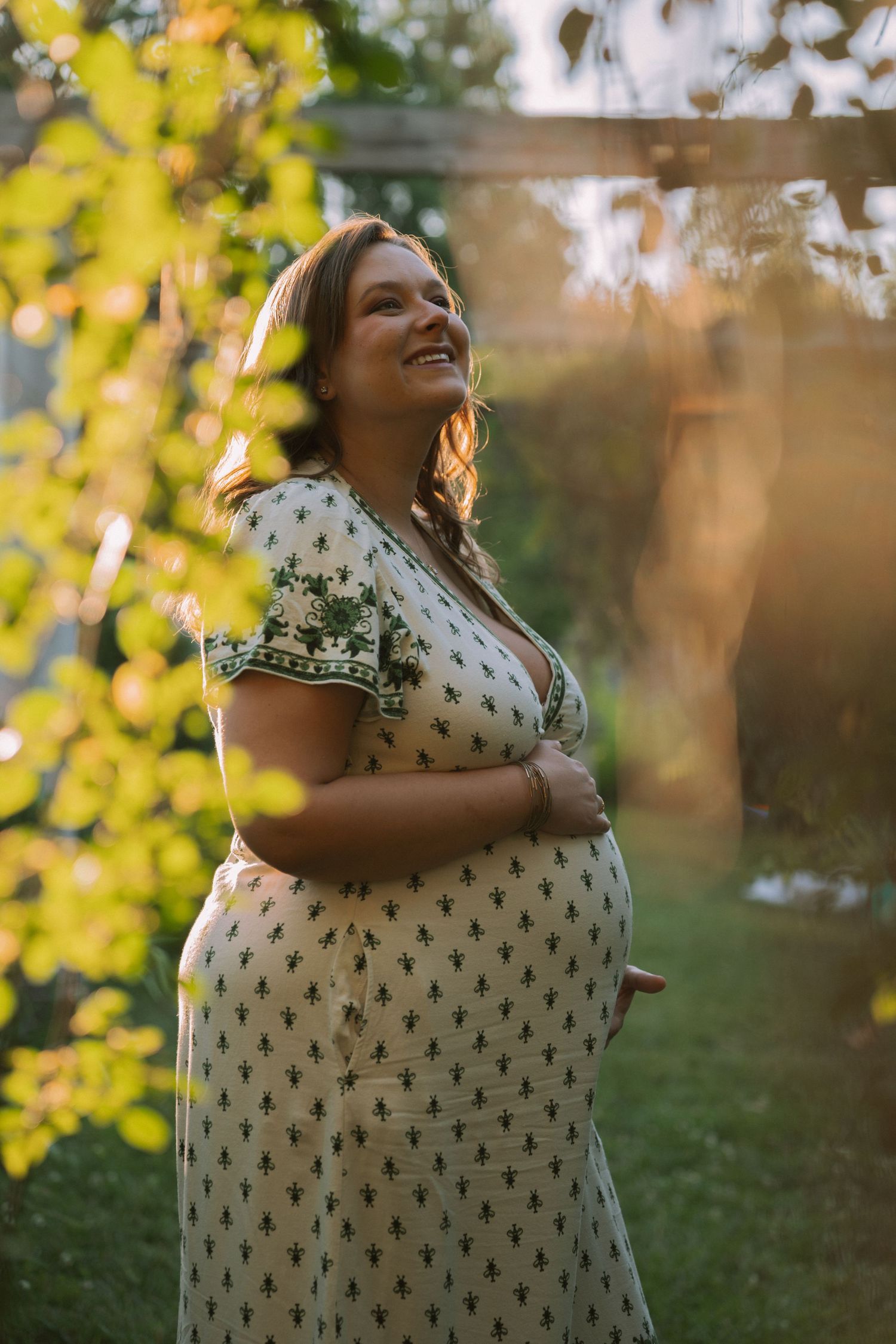 Smiling pregnant woman in floral dress cradling her baby bump outdoors in golden sunlight.