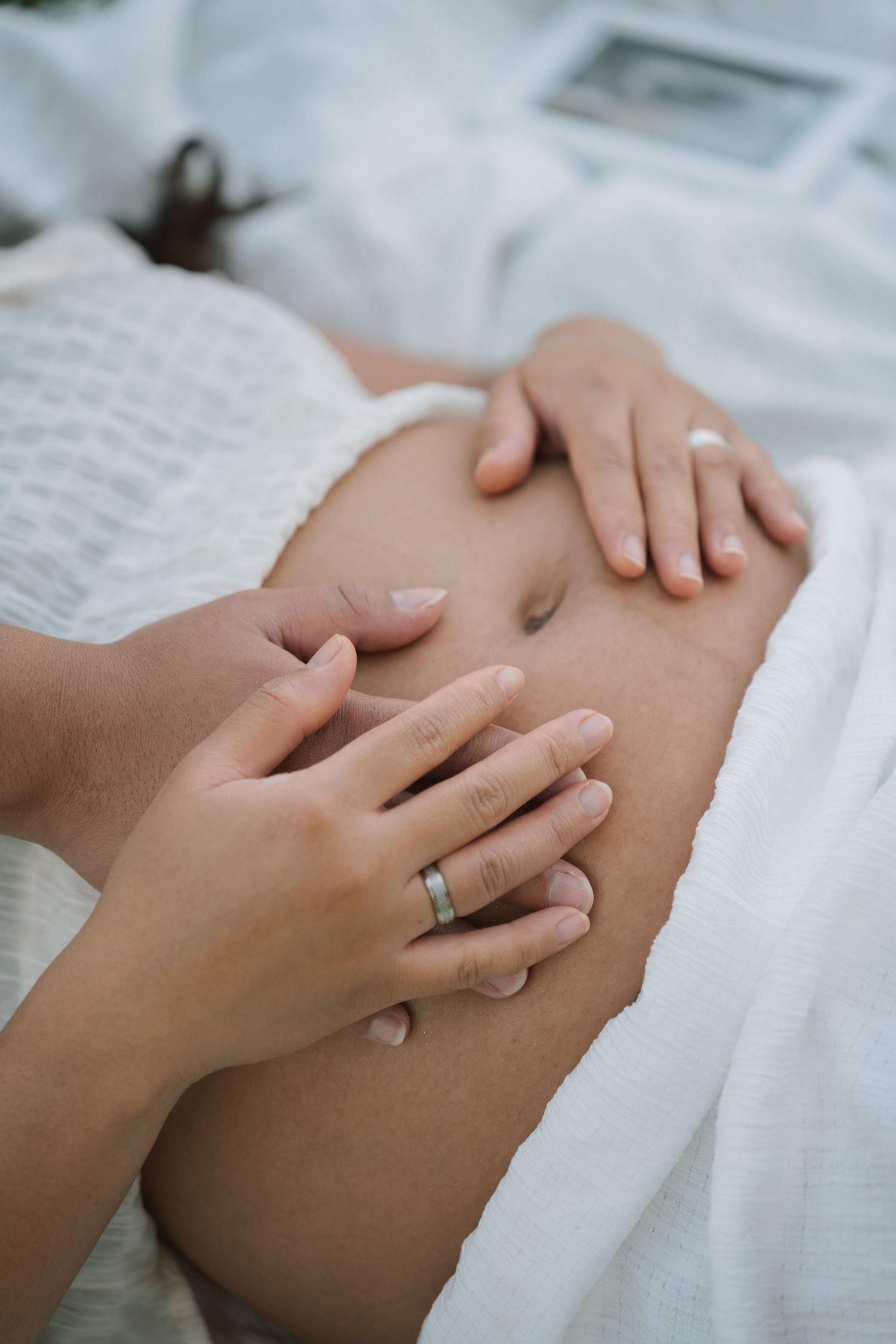 Two hands gently cradling a pregnant belly, with one hand wearing a silver ring, surrounded by soft white fabric.