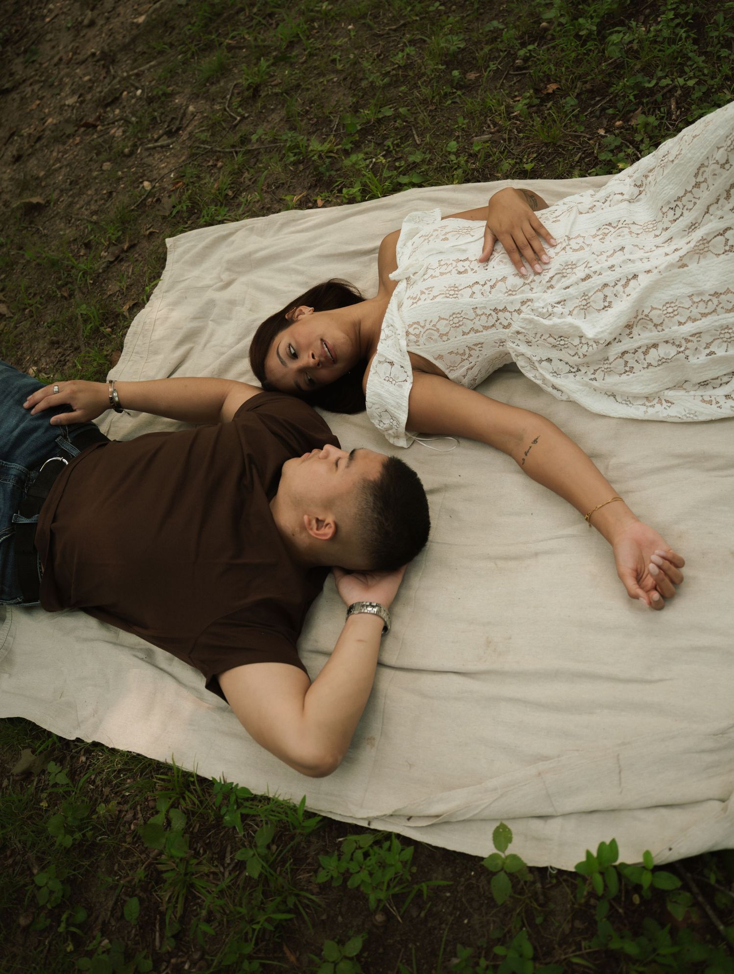 Couple lying on a blanket outdoors, woman in white lace dress touching her baby bump during maternity photo shoot.