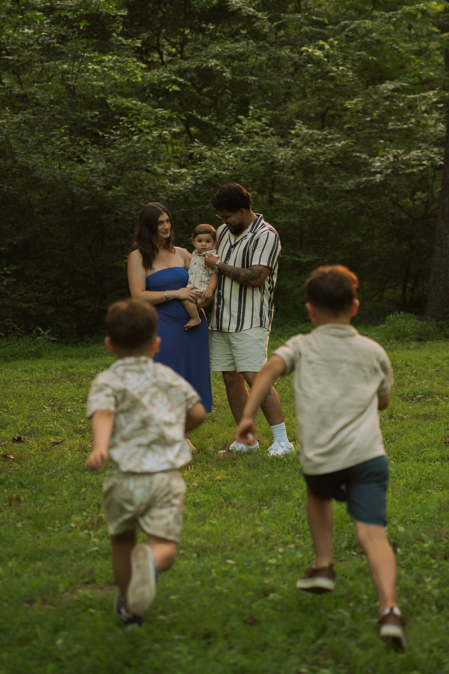 Family of five enjoying a sunny day in a lush green park, with two boys running toward their parents. Row and Co Photography