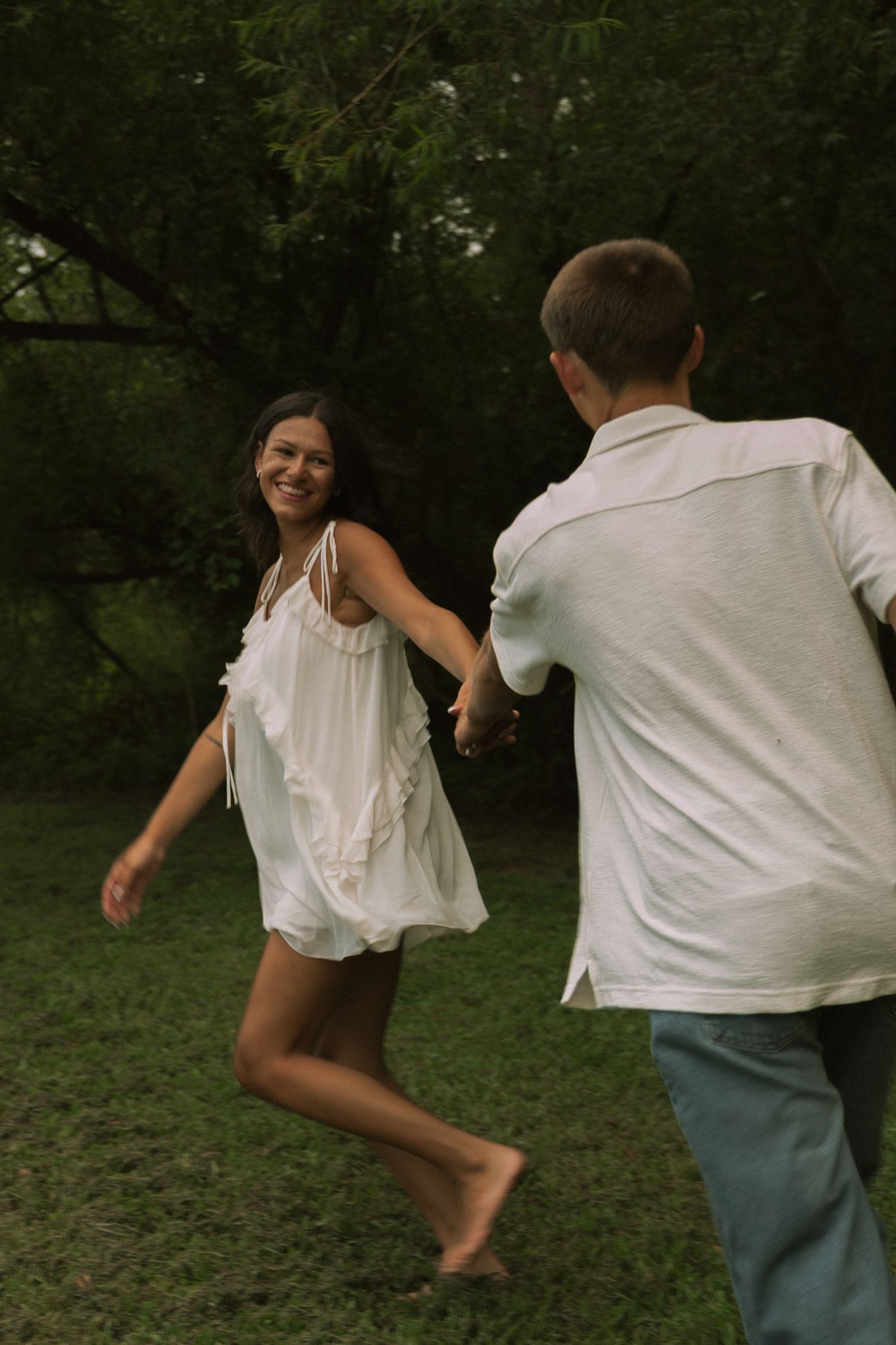 A joyful couple holds hands and dances barefoot on green grass surrounded by lush trees outdoors.