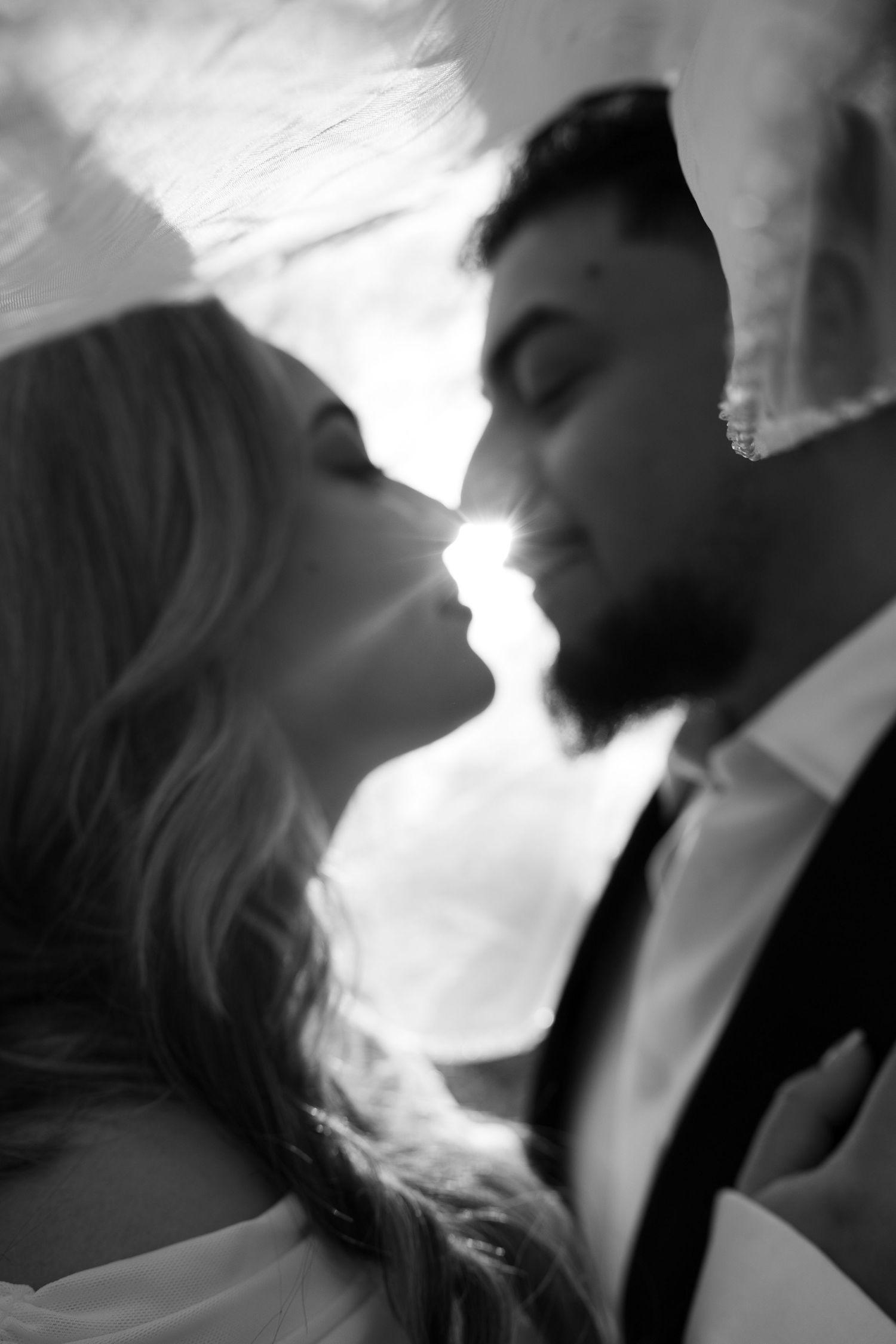 Black and white romantic close-up of bride and groom almost kissing under veil in soft sunlight.