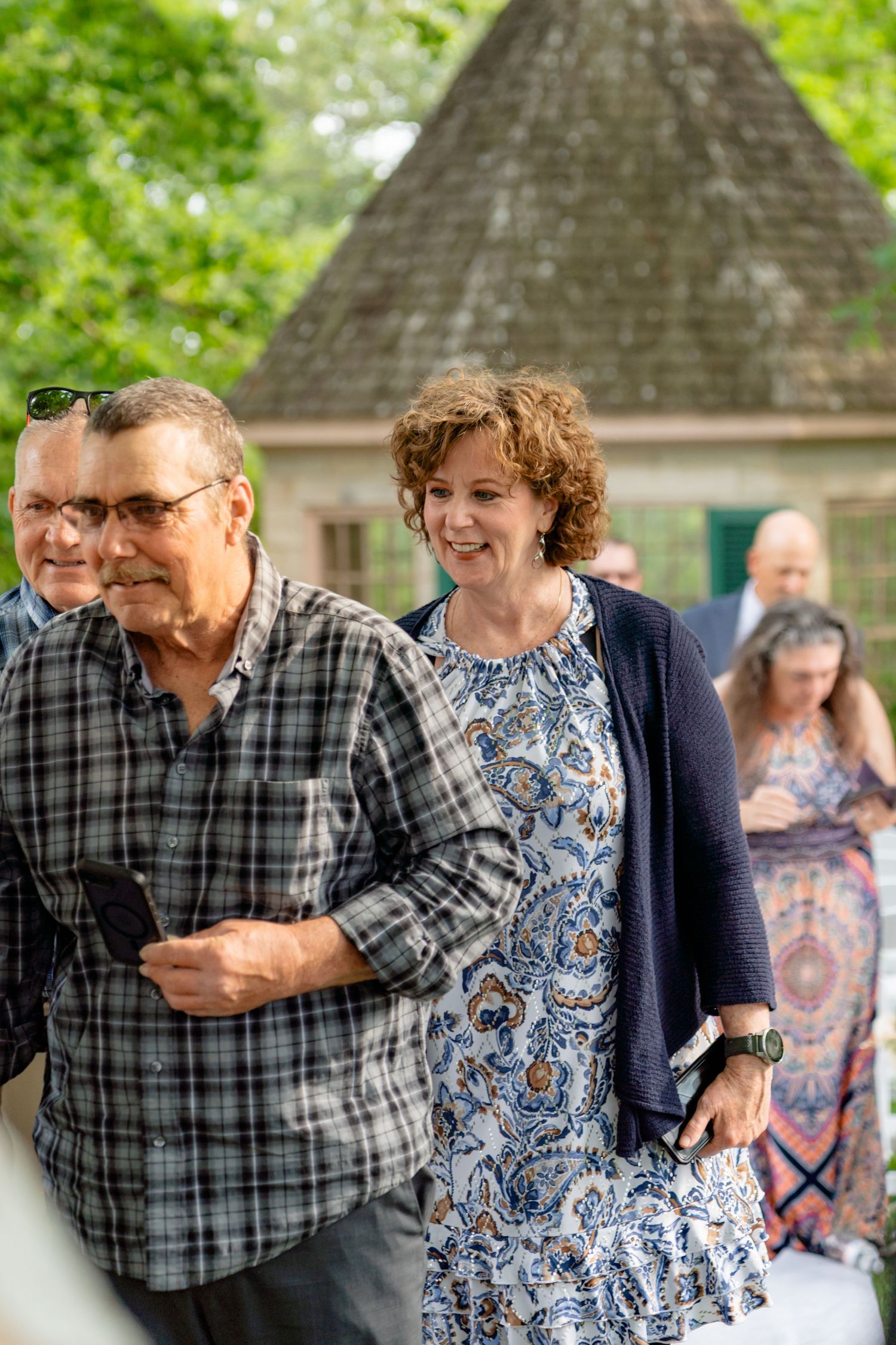 Smiling couple walking outdoors at a garden event, woman in floral dress with navy cardigan, man in plaid shirt.