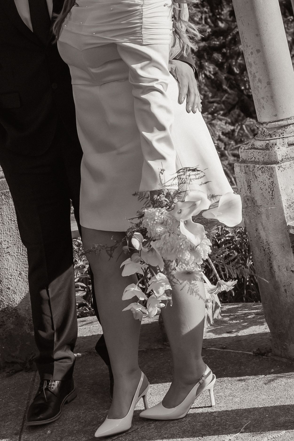 Black and white photo of bride and groom standing together, bride holding bouquet, wearing white dress and heels.