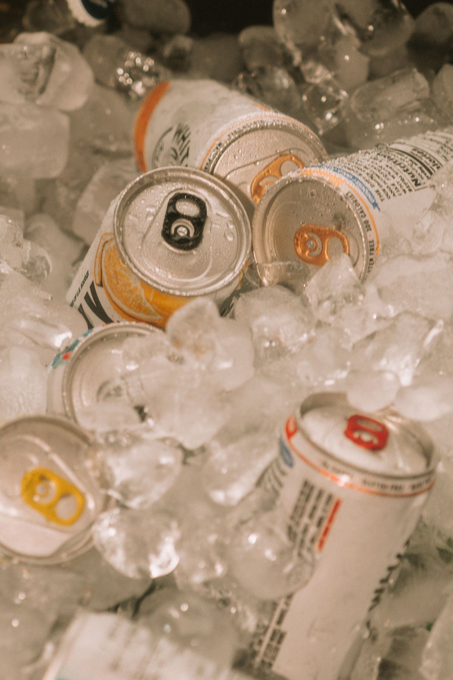 Assorted beverage cans chilling on ice, covered in condensation droplets at a party or event.