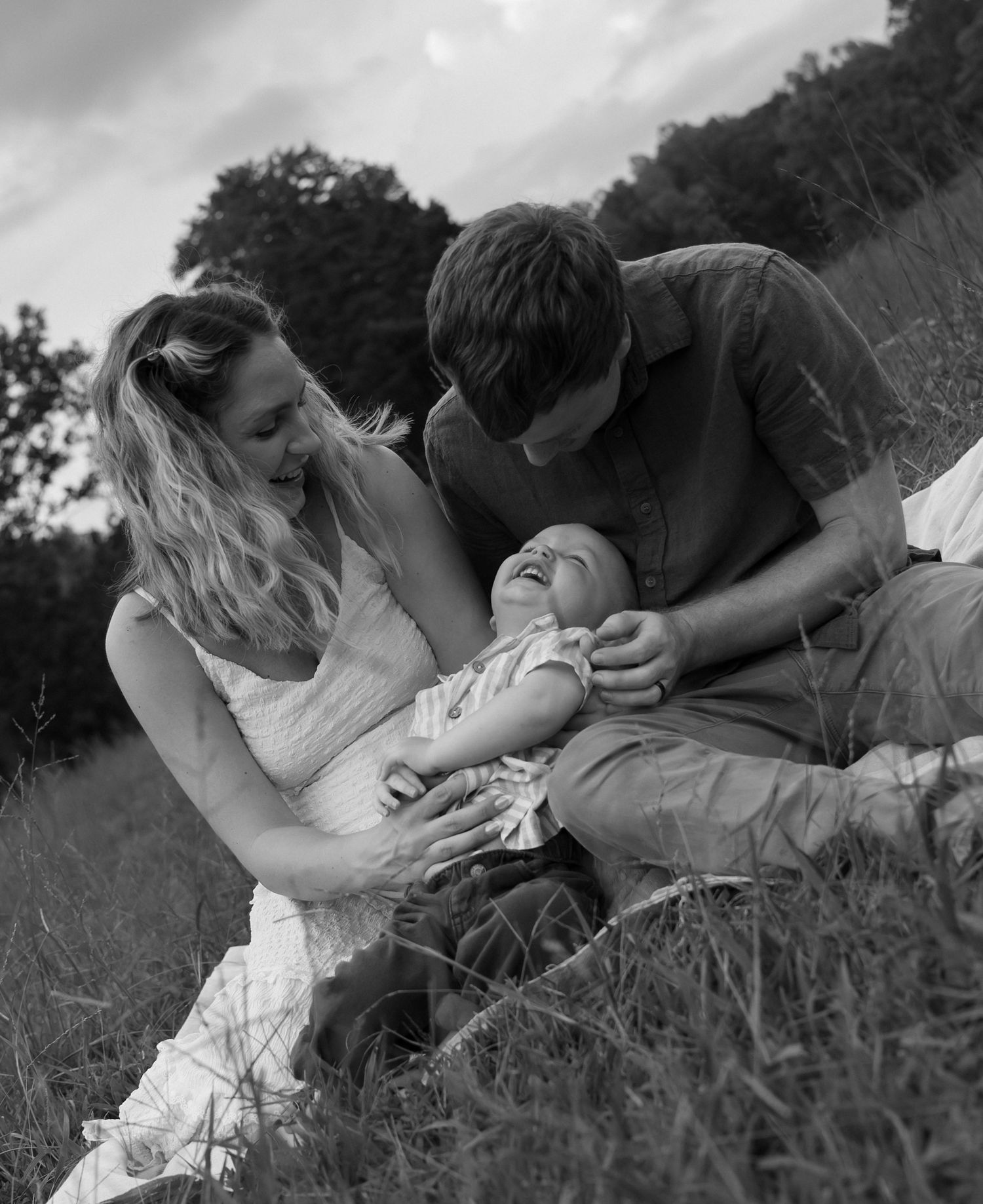 Black and white photo of a happy family with parents laughing with their baby in a grassy field.