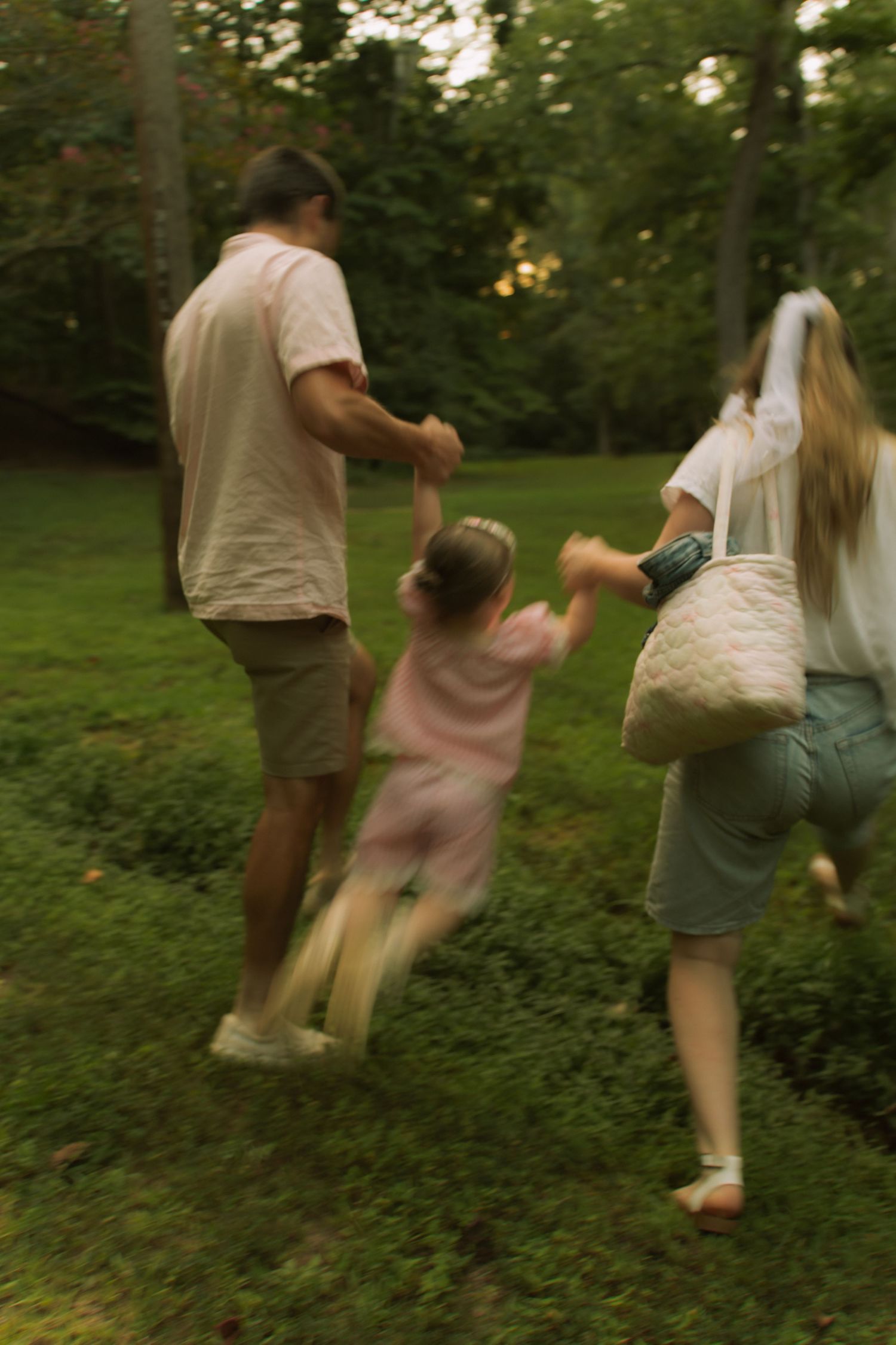 Family walking through lush green park, parents swinging young child between them in motion-blurred outdoor scene.