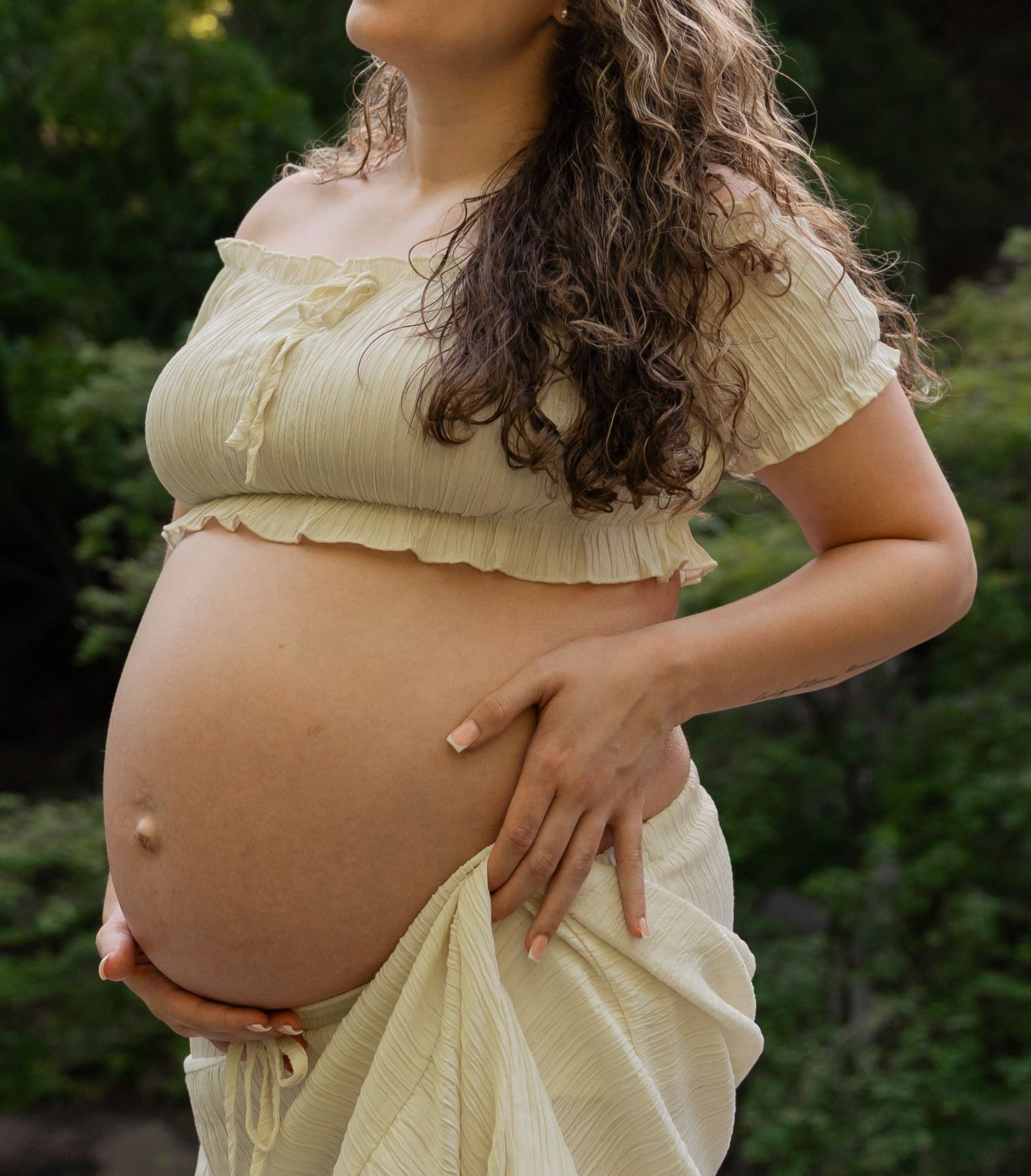 Pregnant woman in yellow crop top and skirt cradling baby bump outdoors with green foliage background.
