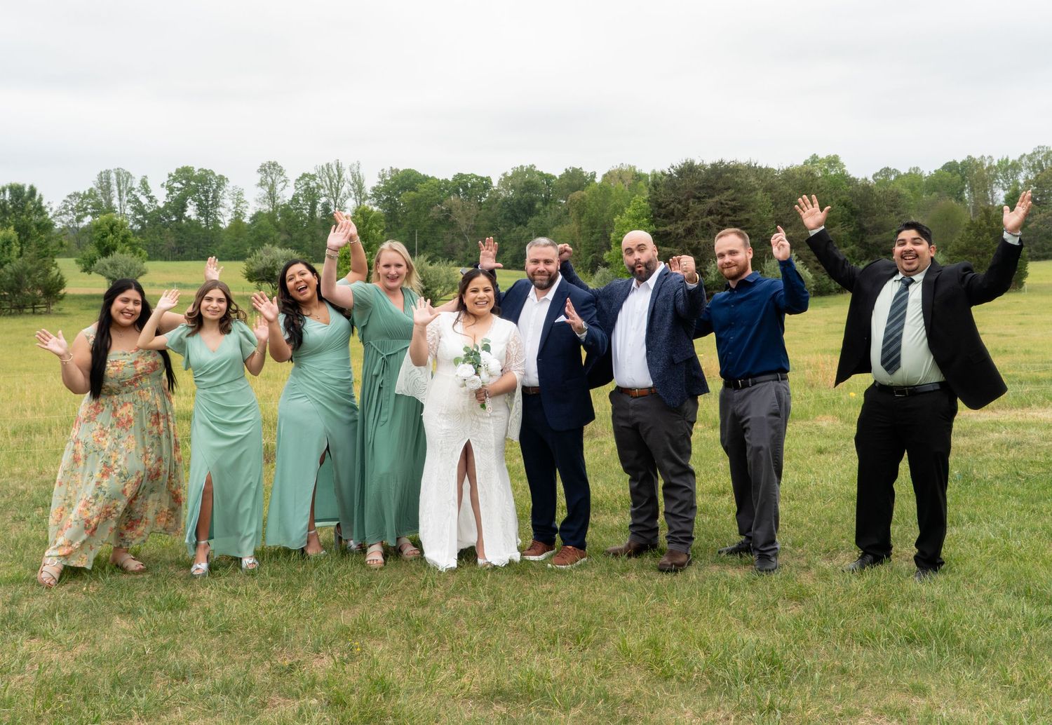 Wedding party celebrating outdoors in a green field, bridesmaids in sage green dresses with cheering guests. Taken by Row And Co Photography