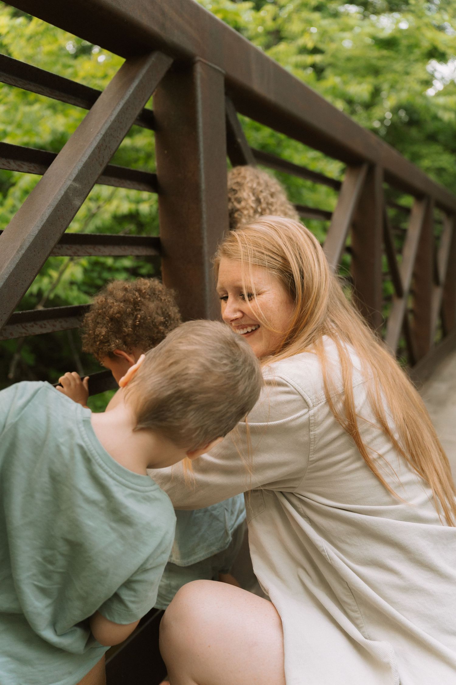 Laughing blonde girl with freckles hugging two boys on a wooden bridge surrounded by green foliage. Taken by Row And Co Photography in FXBG