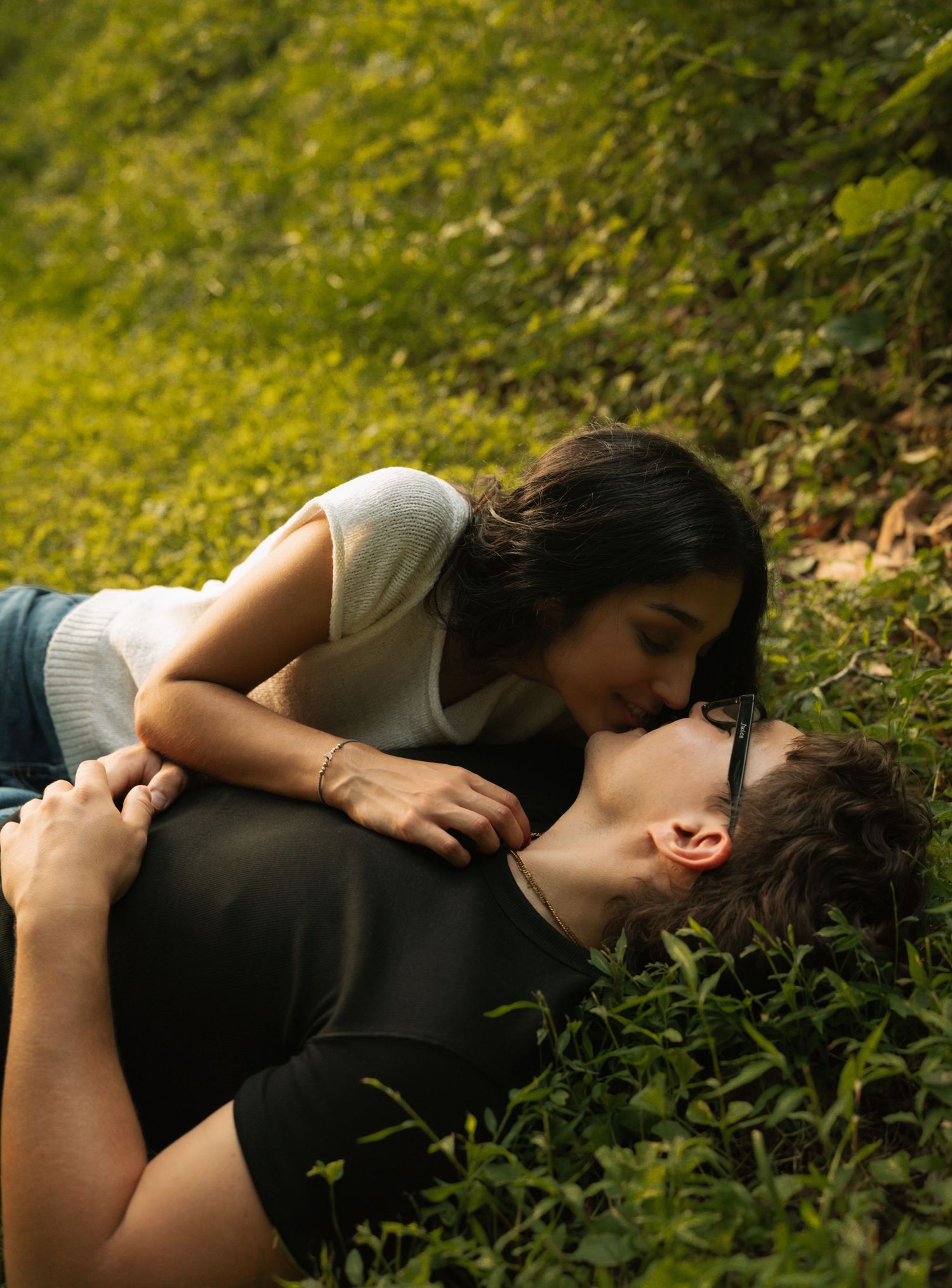 A couple shares a romantic kiss while lying in lush green grass outdoors.