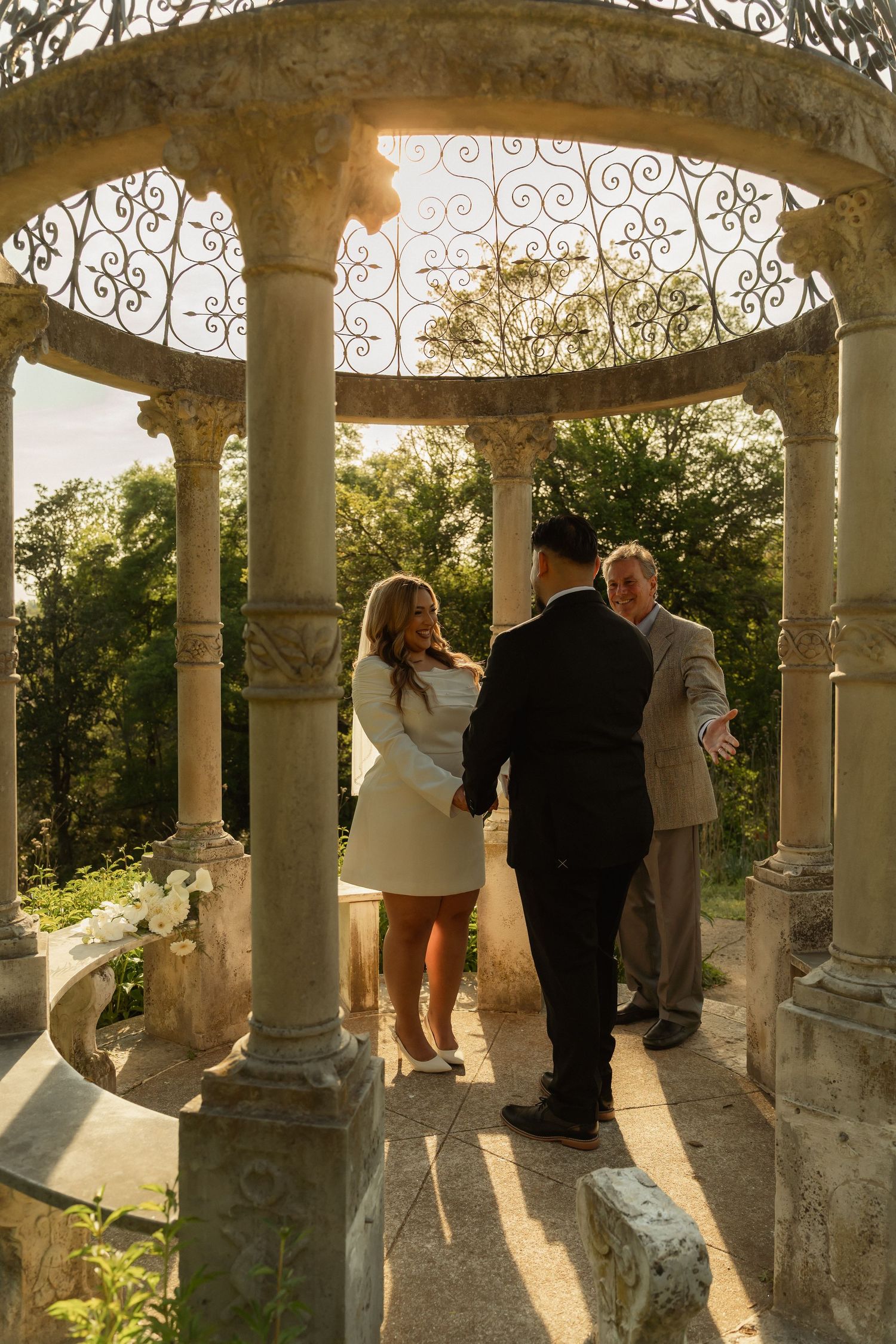 Couple exchanges vows in ornate stone gazebo with wrought iron details, surrounded by lush greenery at golden hour.