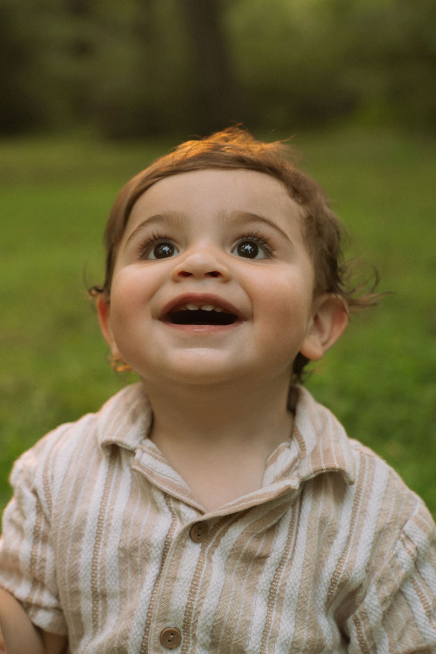 Smiling baby boy with brown eyes wearing a striped shirt, looking up joyfully outdoors in a green park setting.