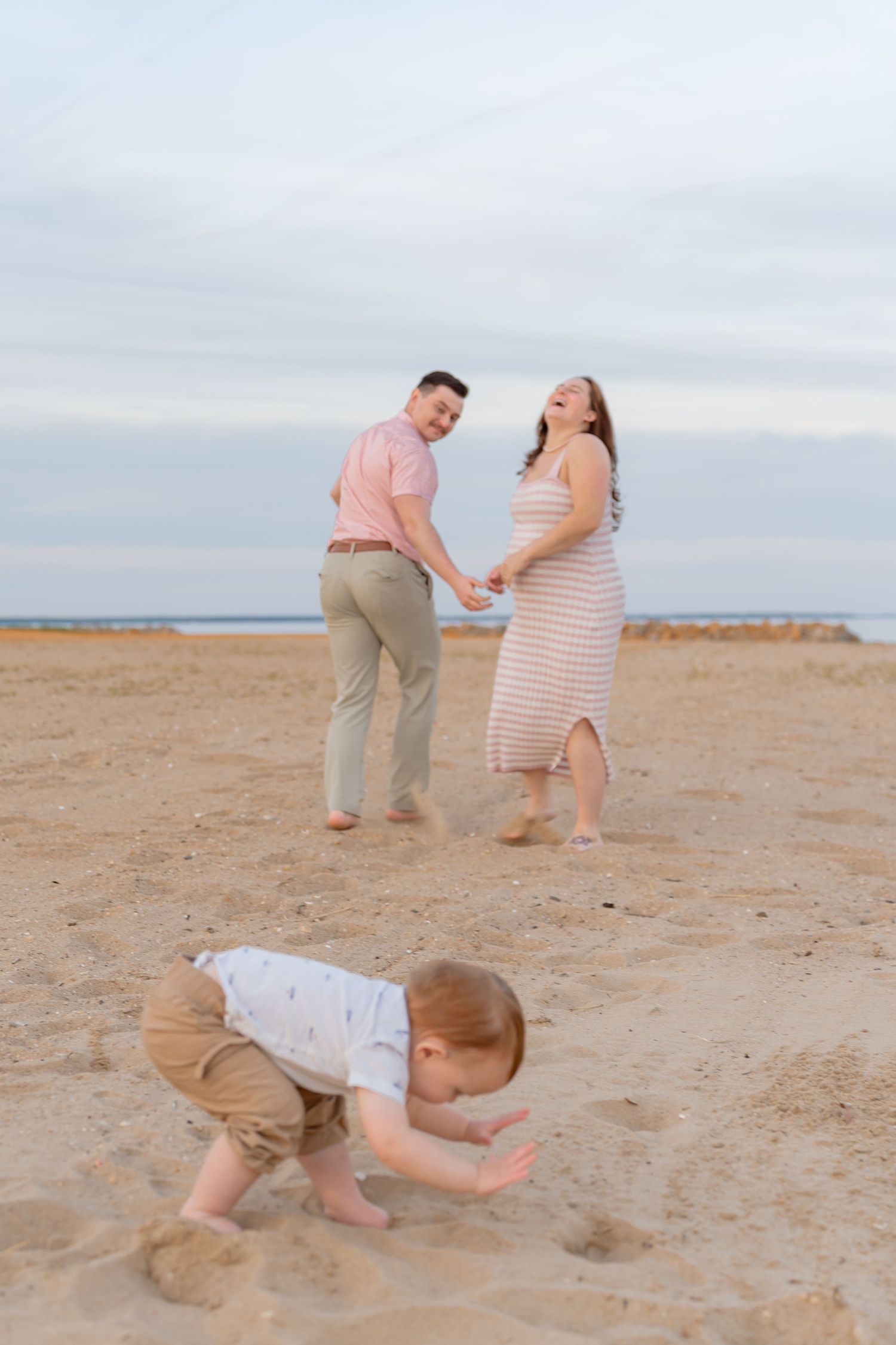 Expecting couple holding hands on a sandy beach while their toddler plays in the sand. Taken by Row And Co Photography in FXBG VA