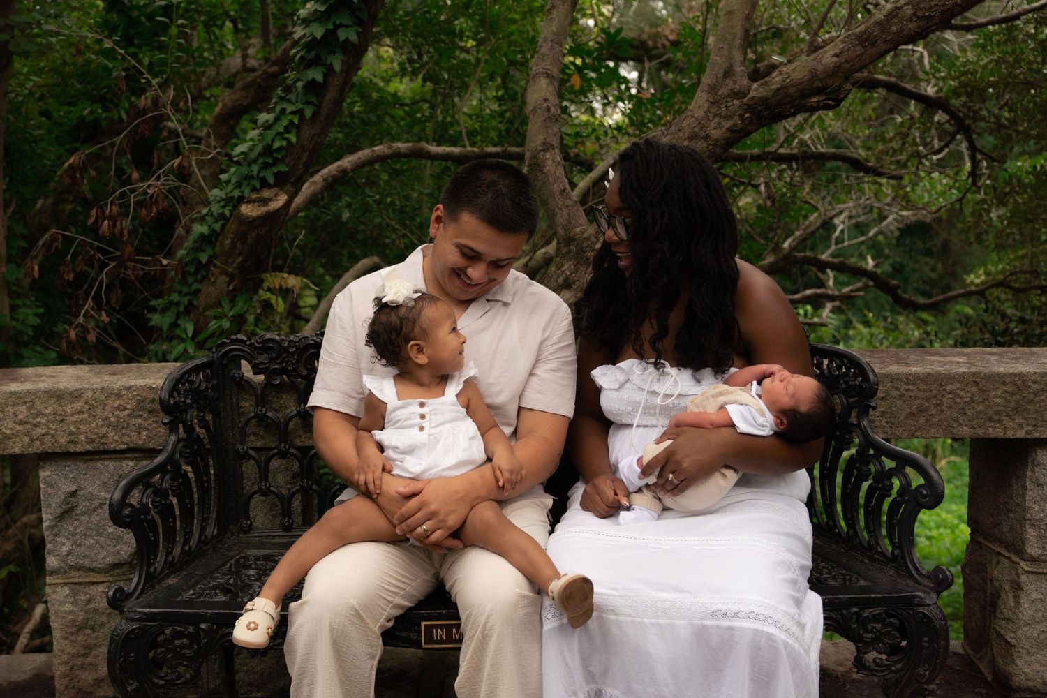 Family of four sitting on ornate bench in lush garden, parents holding two young children in white outfits.