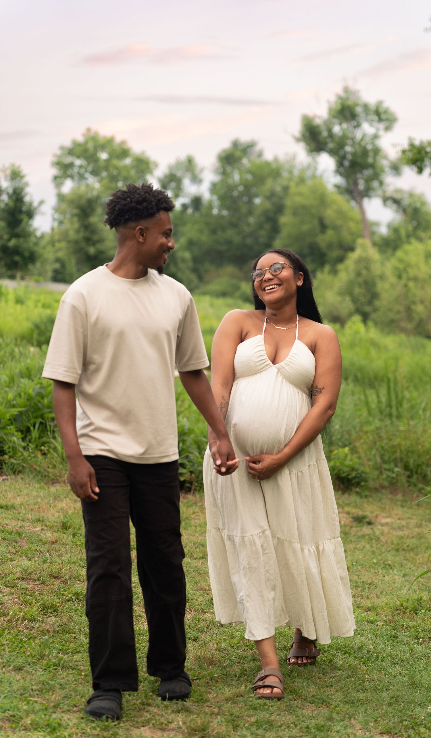 Happy expecting couple holding hands while walking through a lush green field at sunset. Taken by Row And Co Photography in FXBG VA
