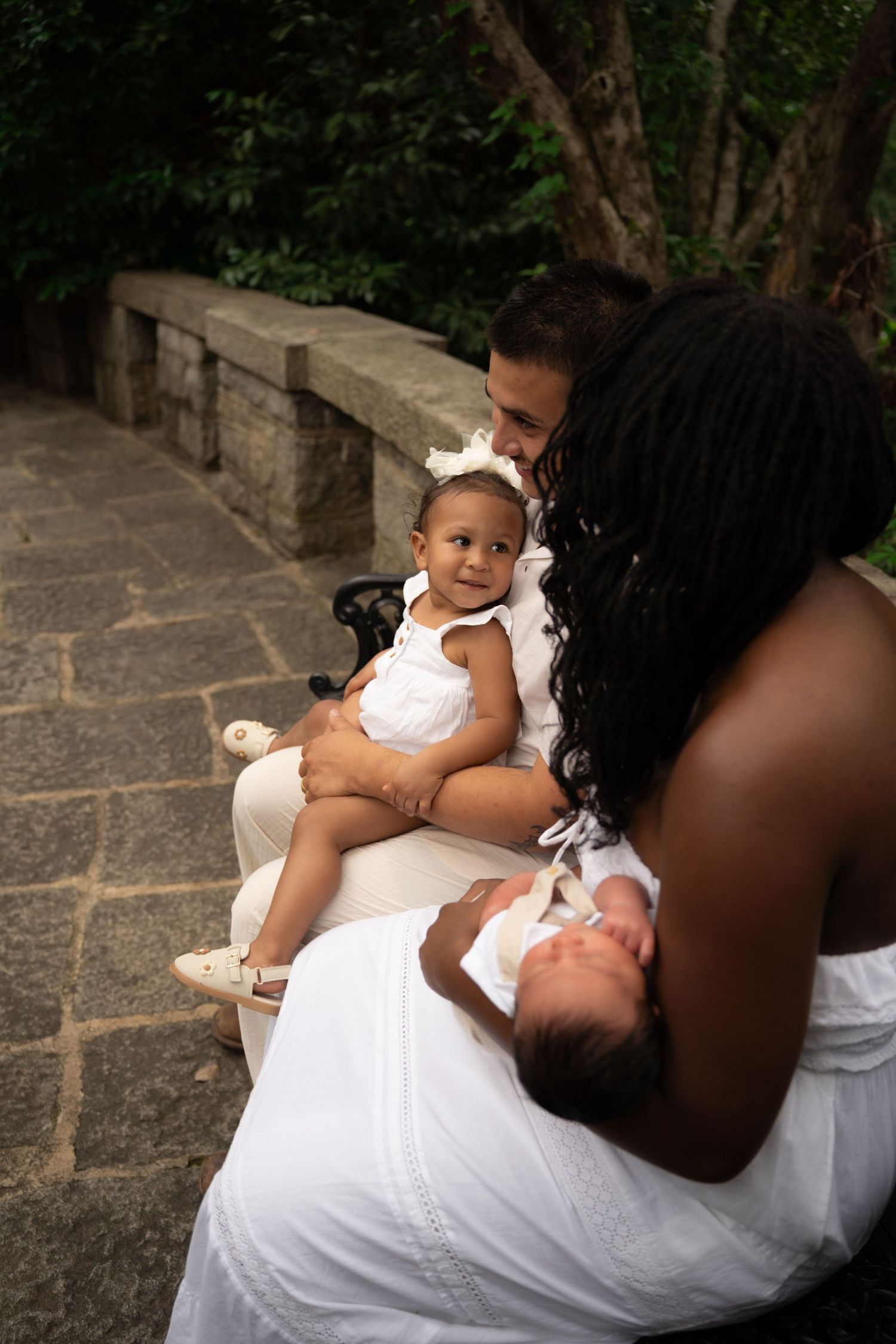 Family sitting outdoors on stone steps, parents in white holding two young children, surrounded by lush greenery.