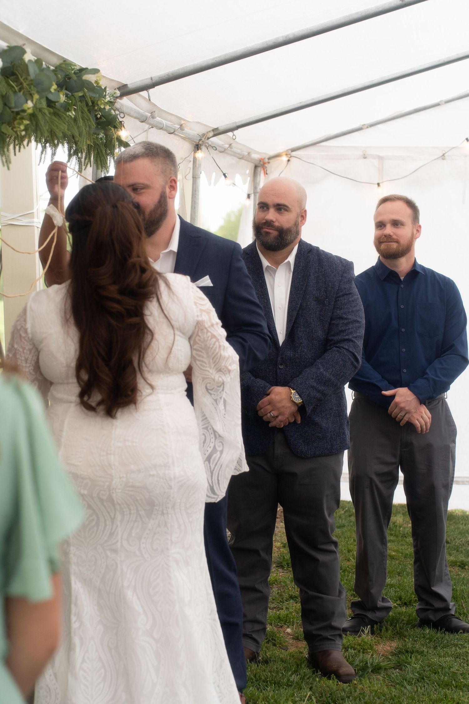 Bride and groom share a kiss at outdoor wedding ceremony under white tent with groomsmen standing nearby.