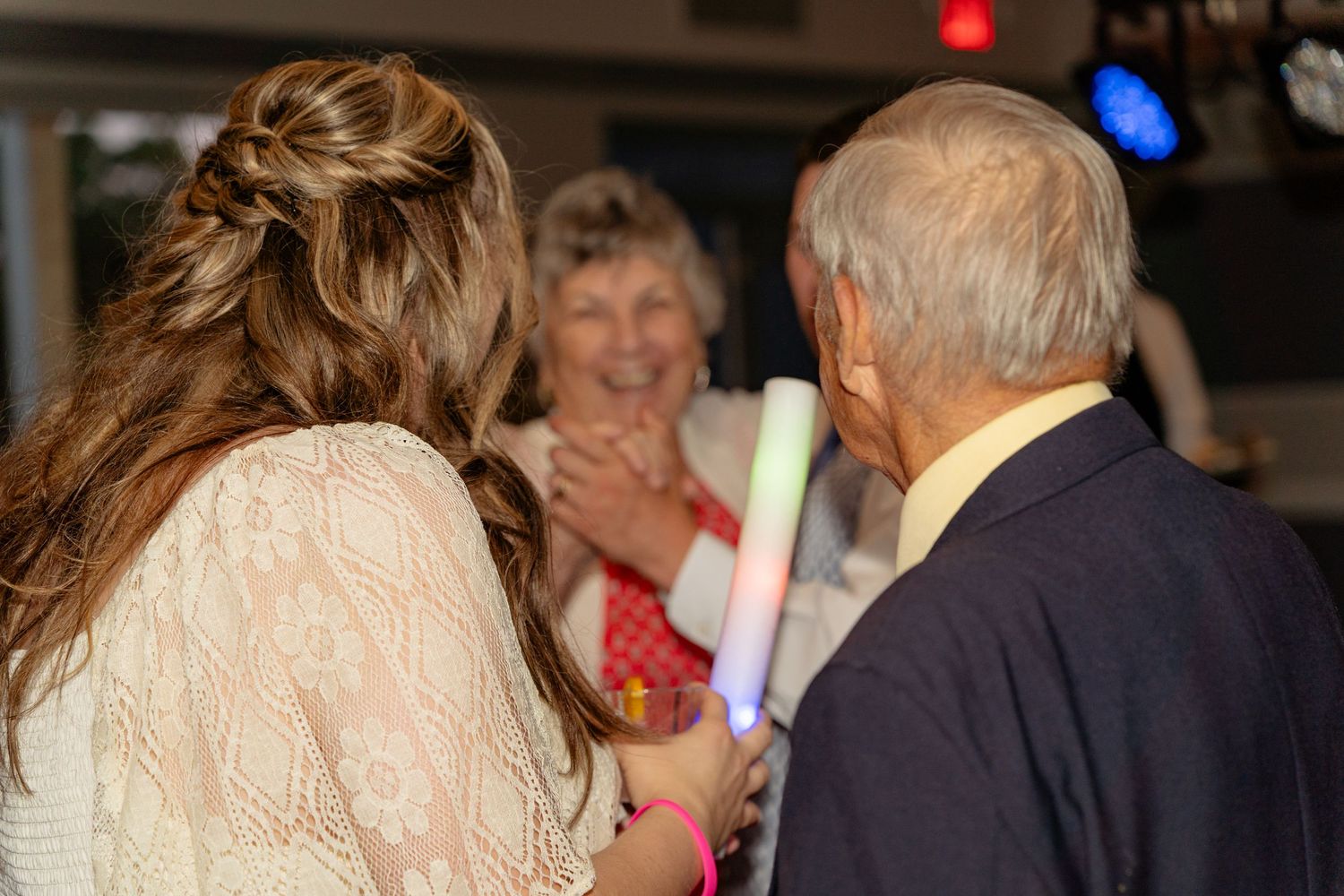 Elderly couple and young woman with braided hair holding glow sticks at a wedding reception.