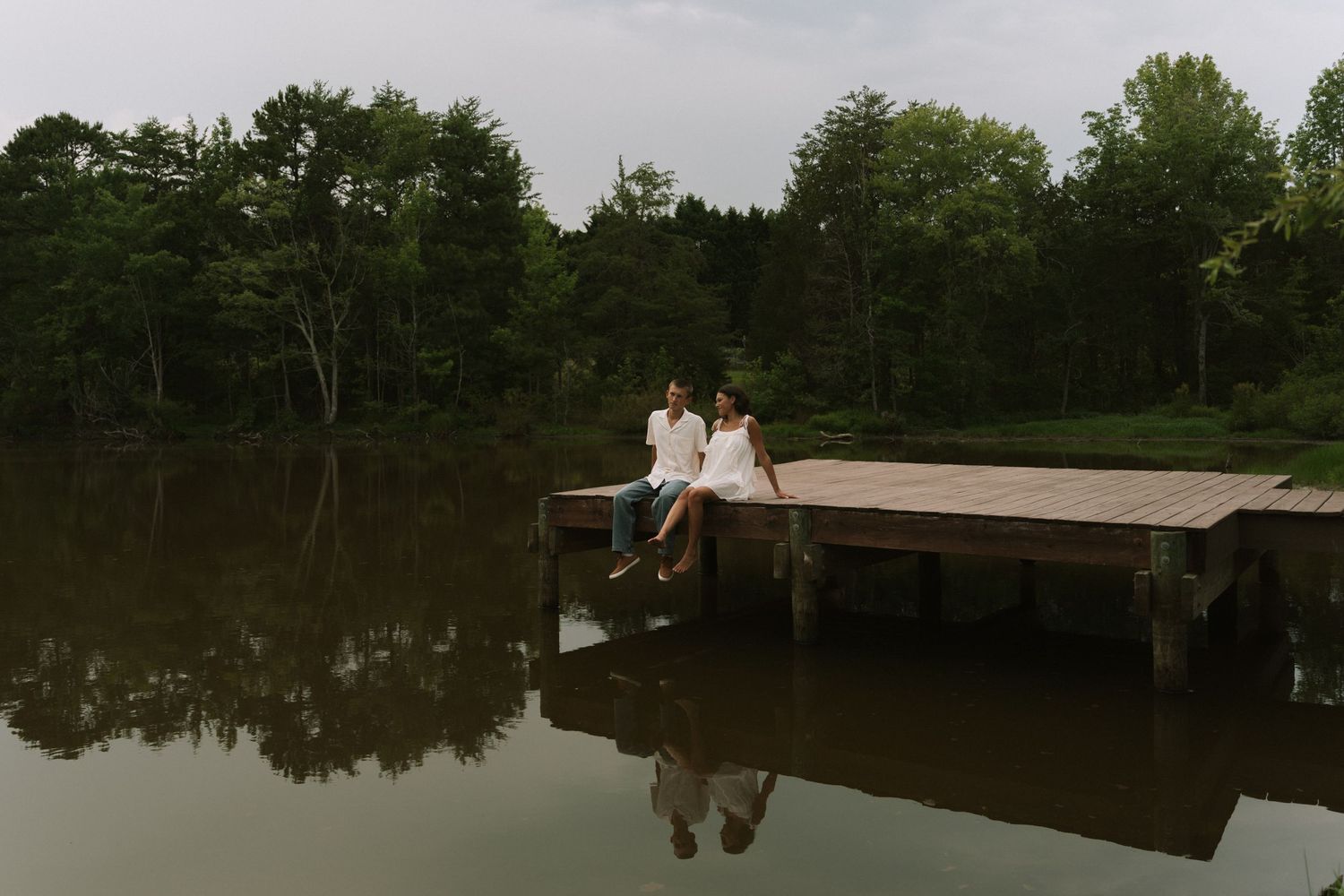 Couple sitting together on a wooden dock over a calm lake surrounded by lush green trees.