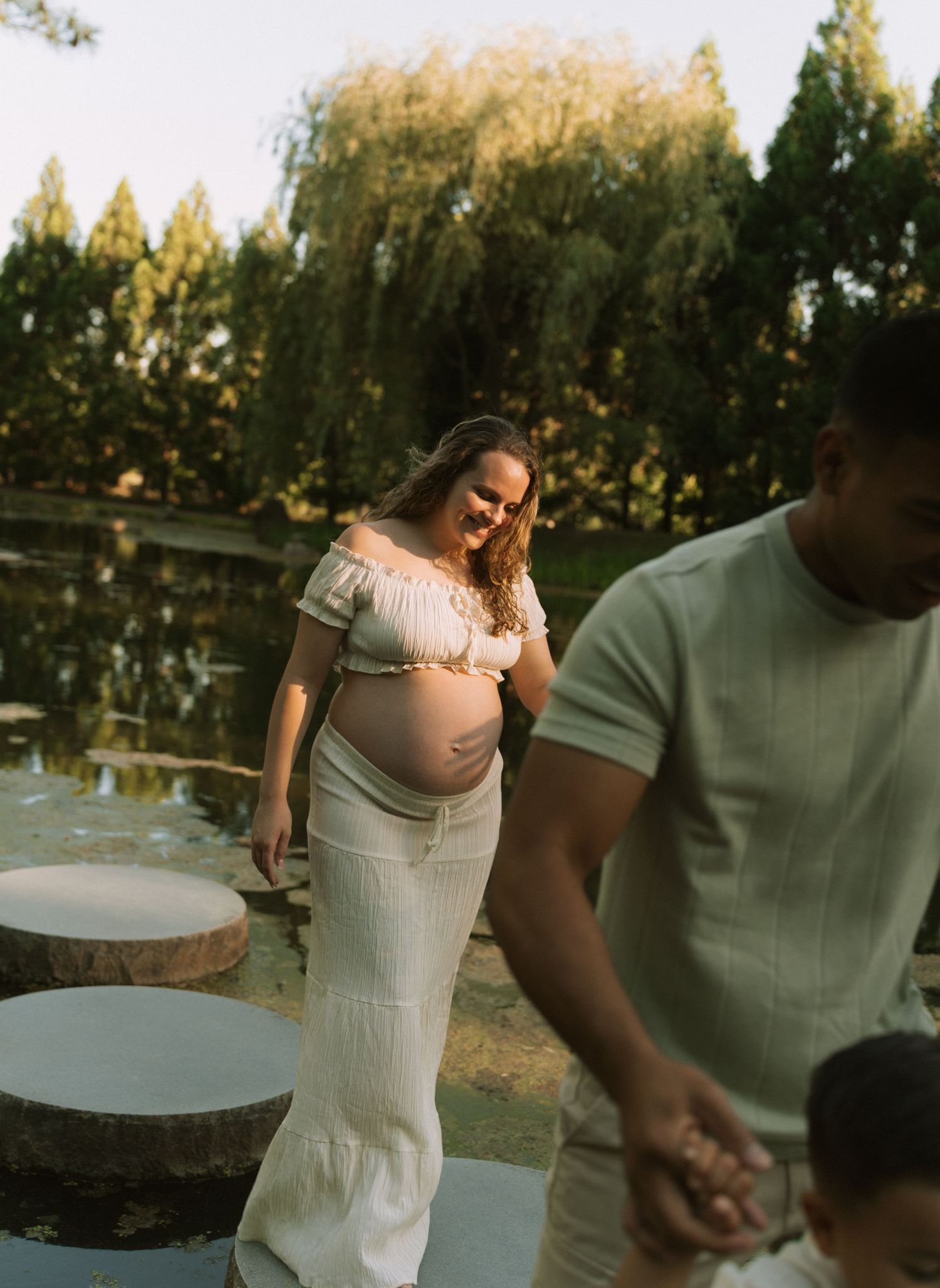 Pregnant woman in white skirt and crop top smiling outdoors near a pond with a man and child.