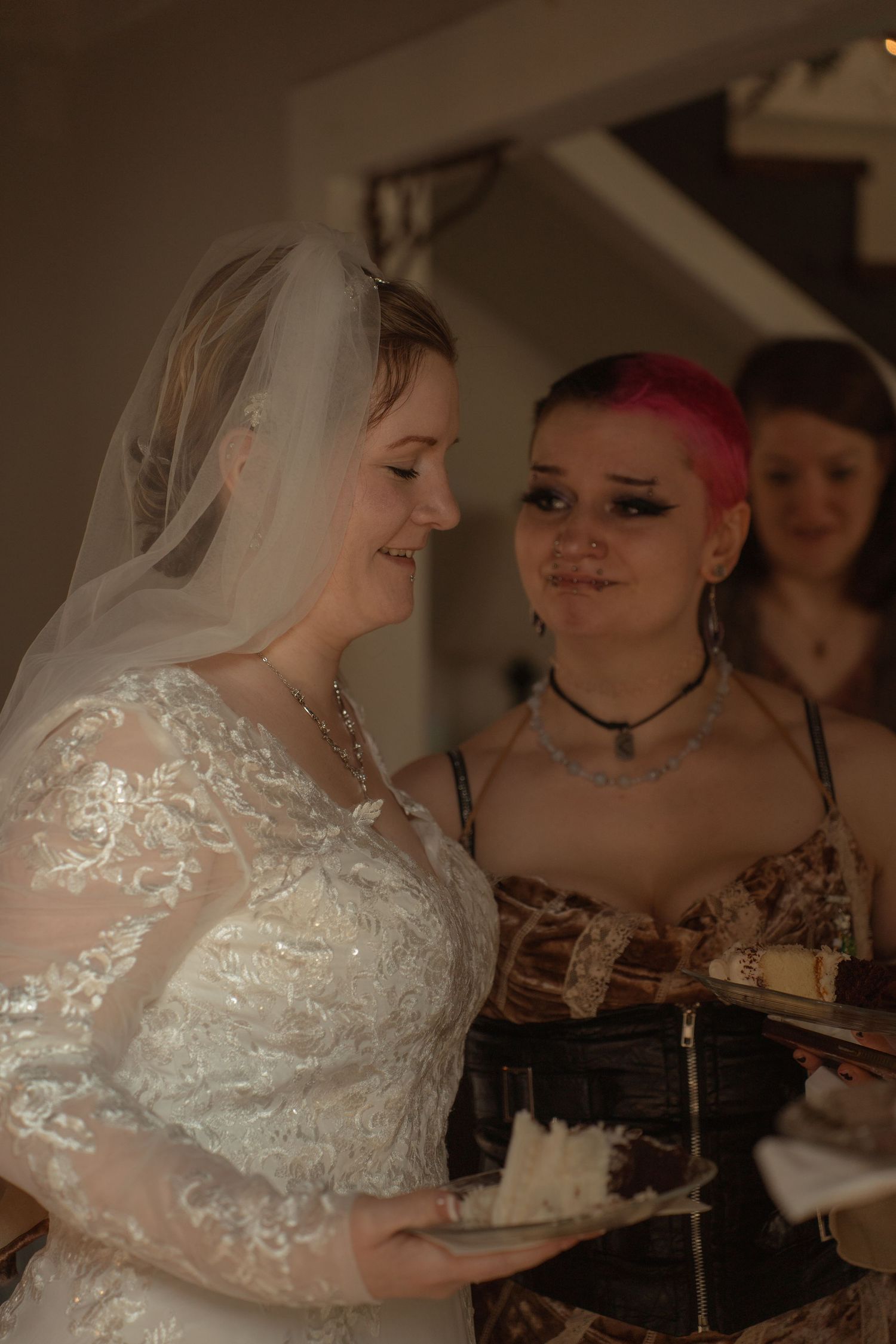 Bride in lace wedding dress and veil smiles beside pink-haired bridesmaid in corset dress during indoor wedding ceremony.