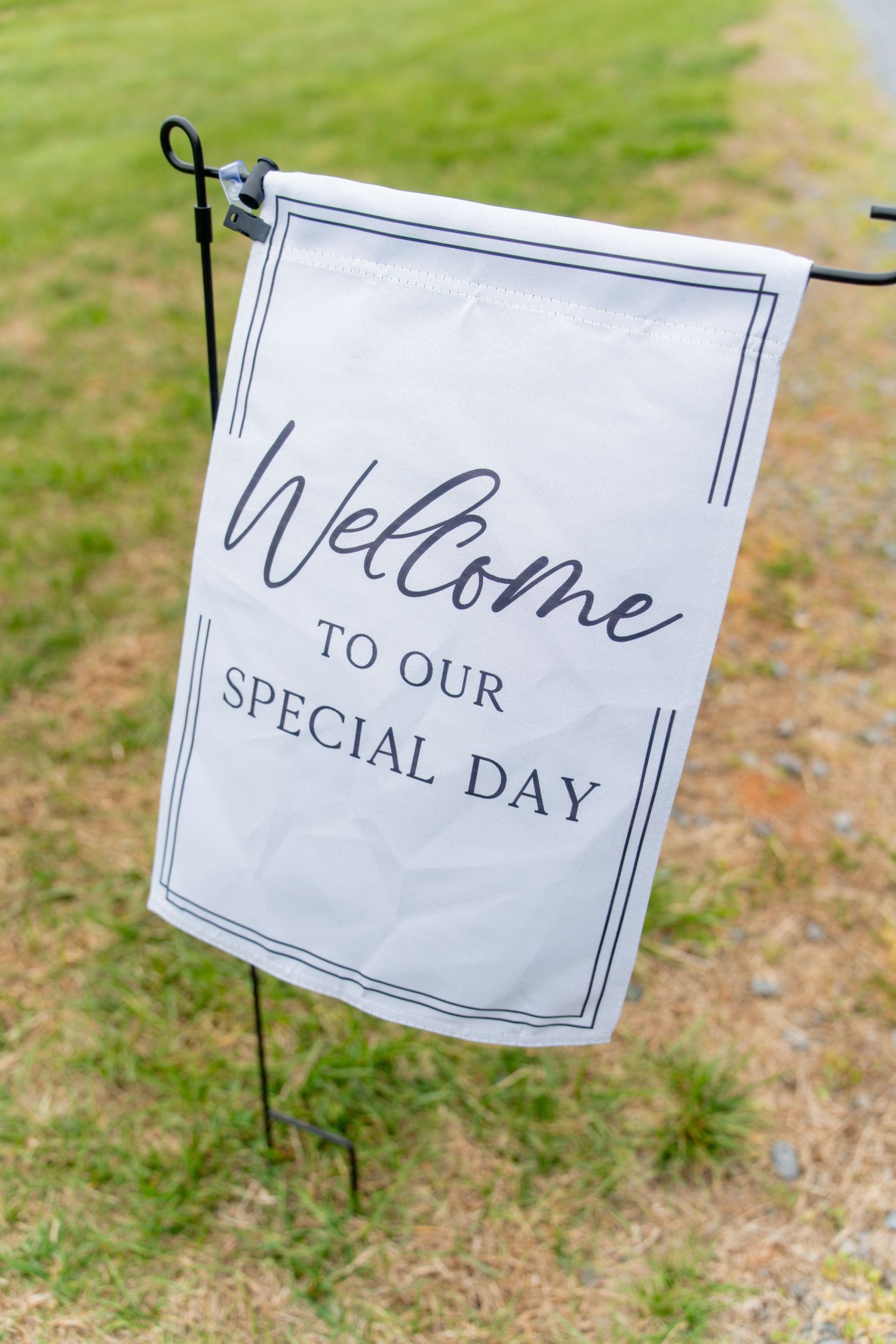 White garden flag on metal stake reading Welcome to Our Special Day, displayed on grass at an outdoor wedding.