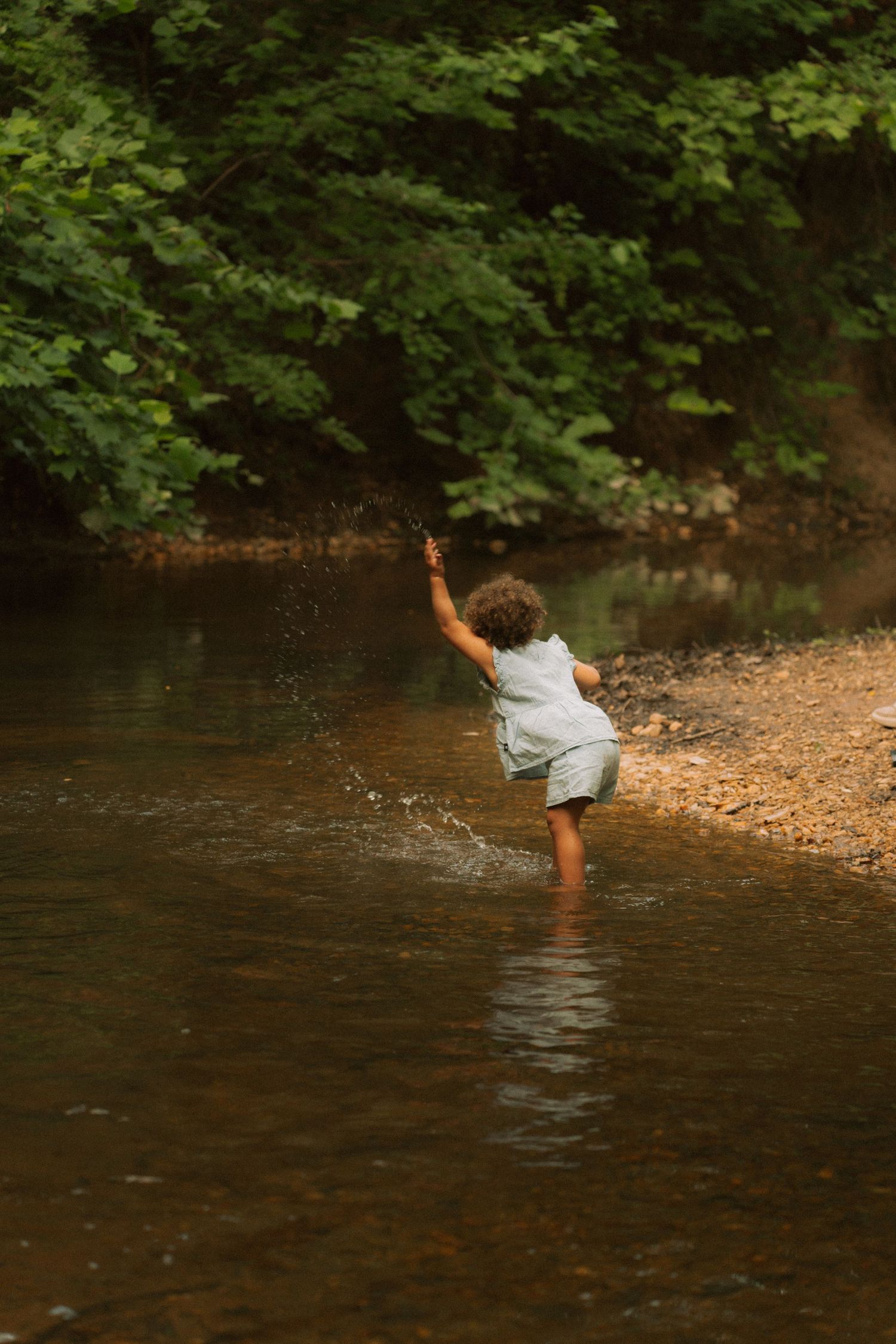 A curly-haired child splashes and plays in a shallow creek surrounded by lush green trees. Taken by Row And Co Photography in FXBG VA