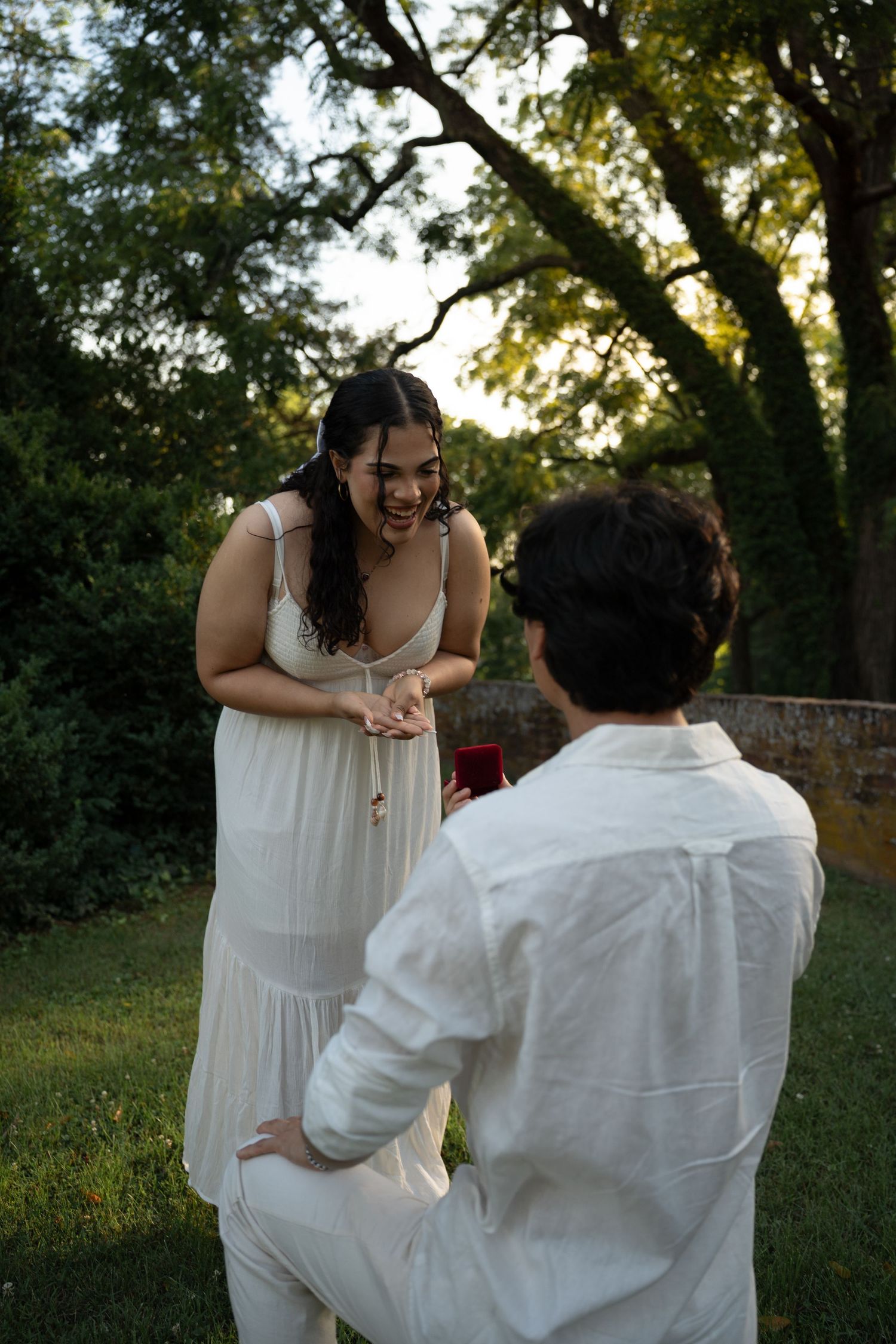 Joyful marriage proposal in a lush garden, man on one knee presenting ring box to laughing woman. Taken by Row And Co Photography in FXBG