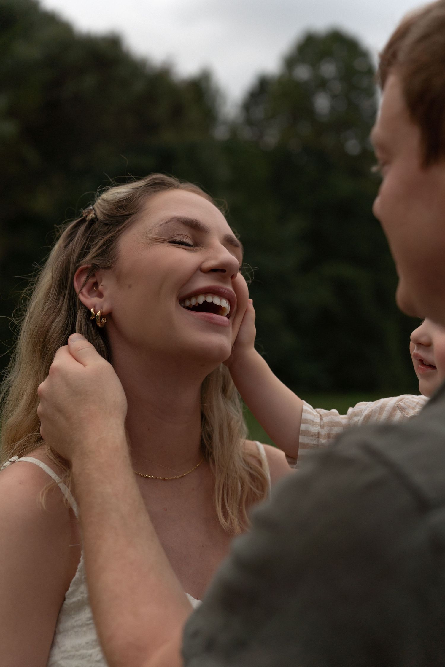 A joyful woman laughs as a man gently touches her face outdoors with lush green trees in the background.