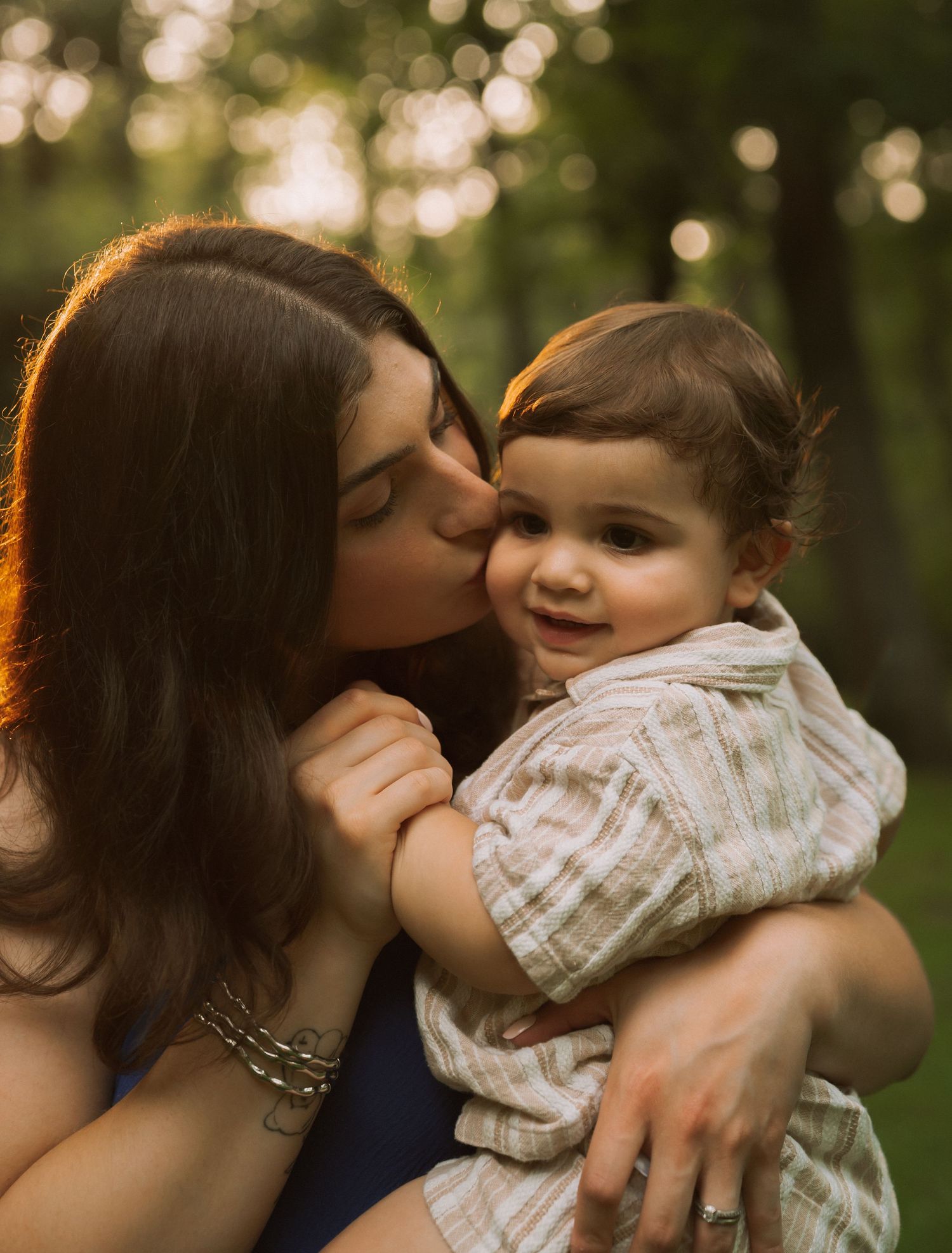 A mother tenderly kisses her smiling toddler outdoors in golden hour light.