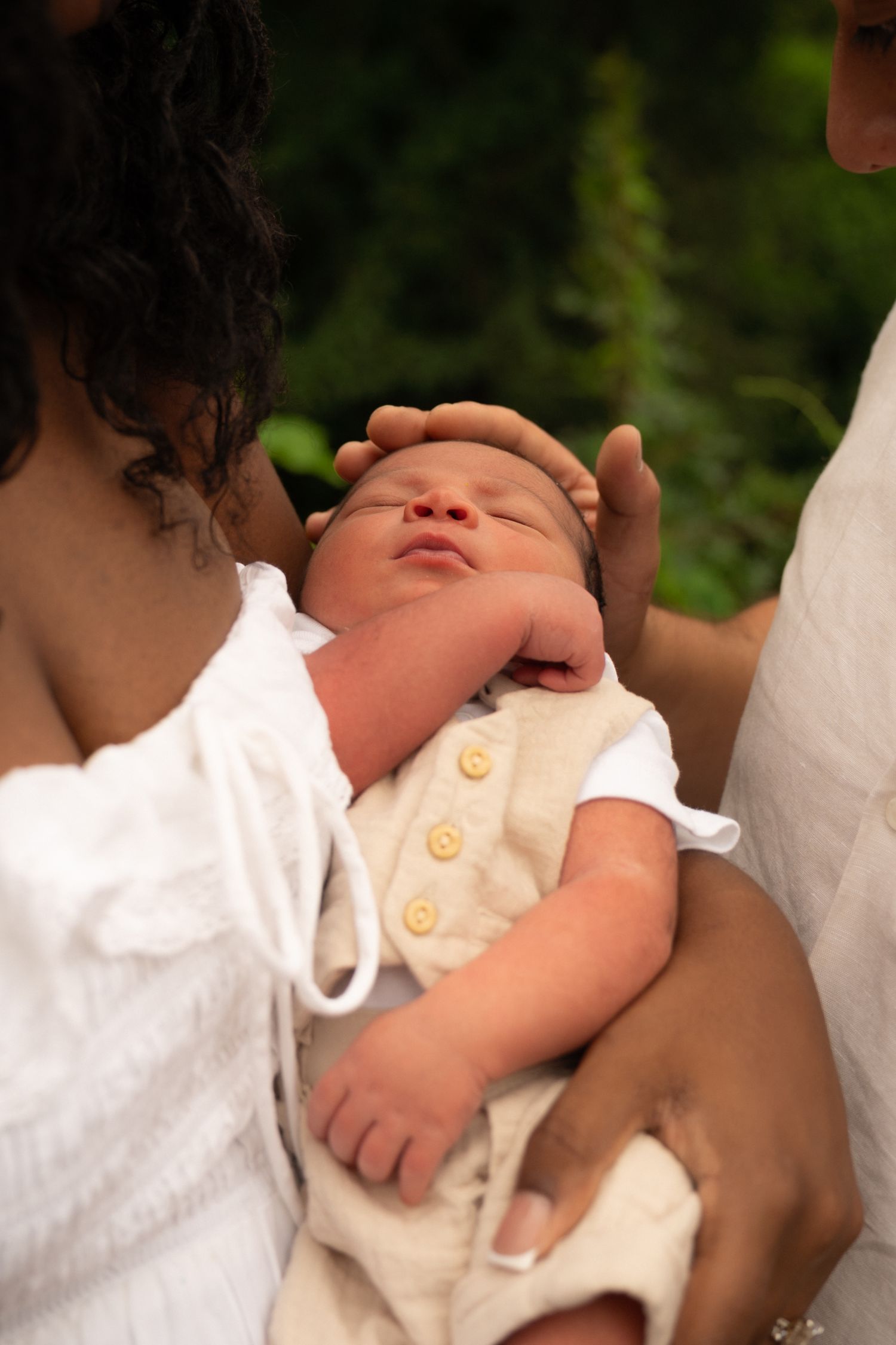 Newborn baby in beige vest with yellow buttons, cradled in arms of two adults wearing white outdoors.