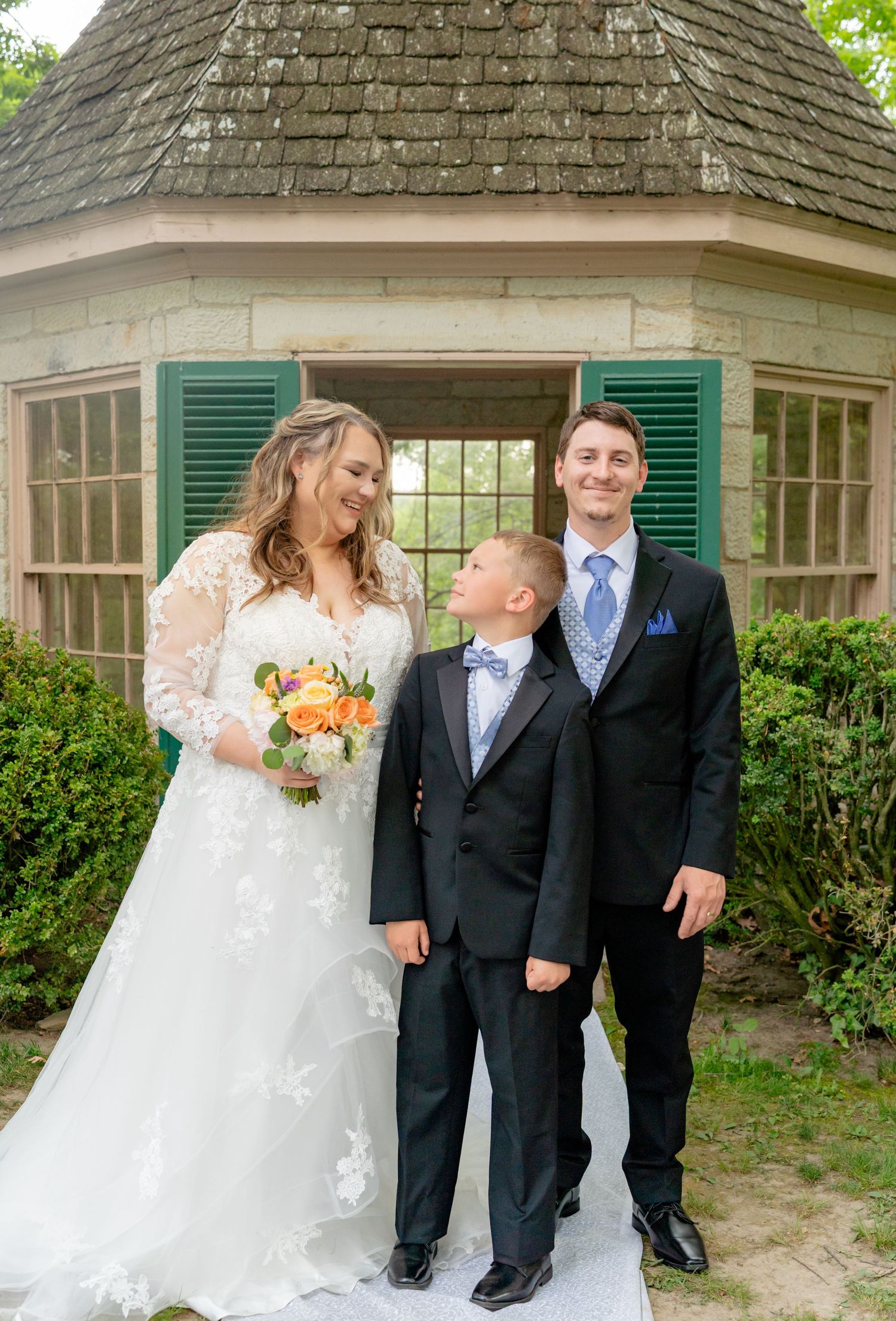 Bride in lace gown holds bouquet beside groom in black suit and young boy in tuxedo at outdoor gazebo wedding. Row and Co Photography