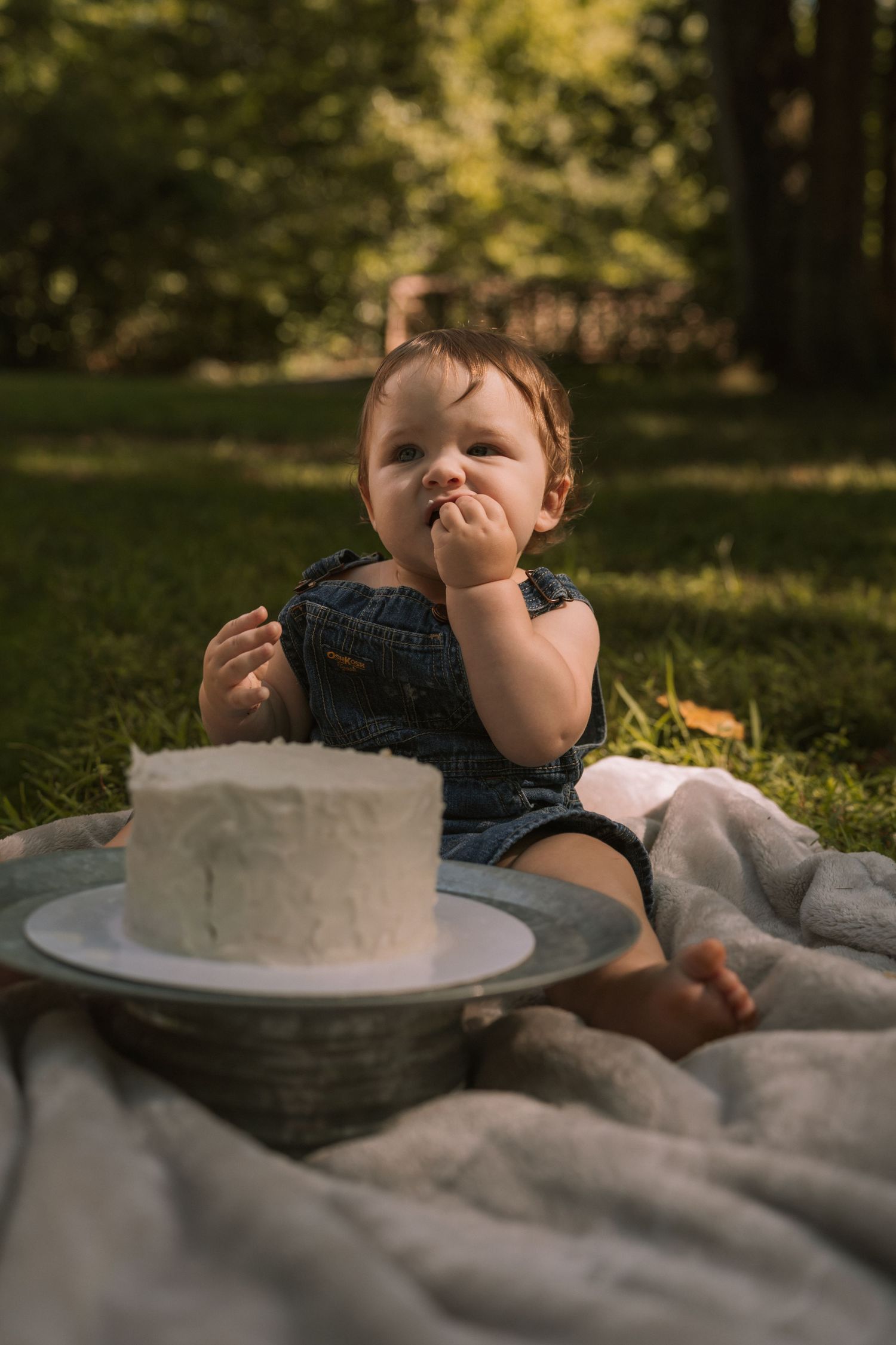 Baby in denim romper sitting on blanket in grass, eating white frosting from first birthday smash cake outdoors.