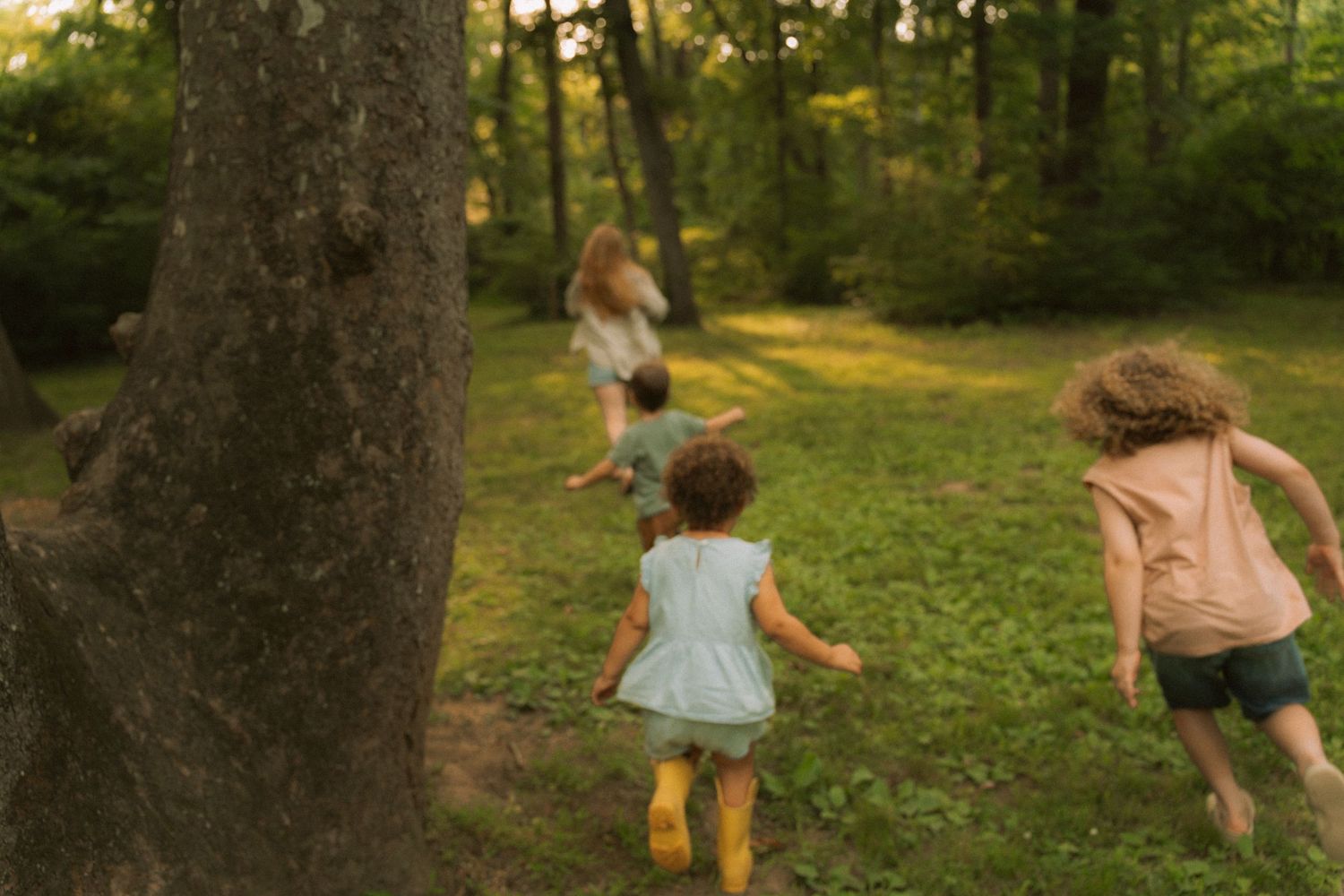 Three young children running through sunlit green woods during outdoor family session. Taken by Row And Co Photography in FXBG VA