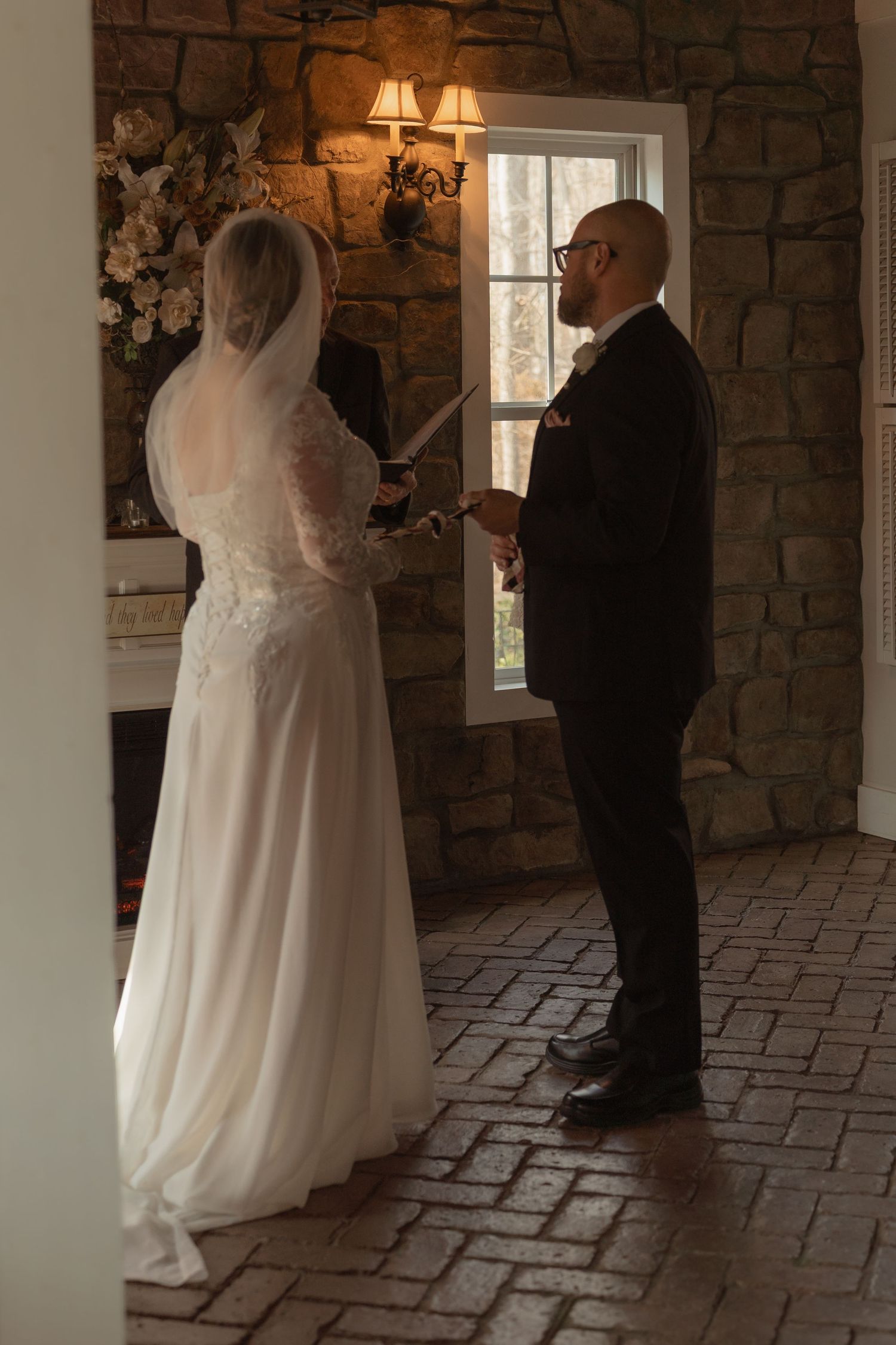 Bride and groom exchange vows indoors by a stone wall and window in a romantic wedding ceremony setting.