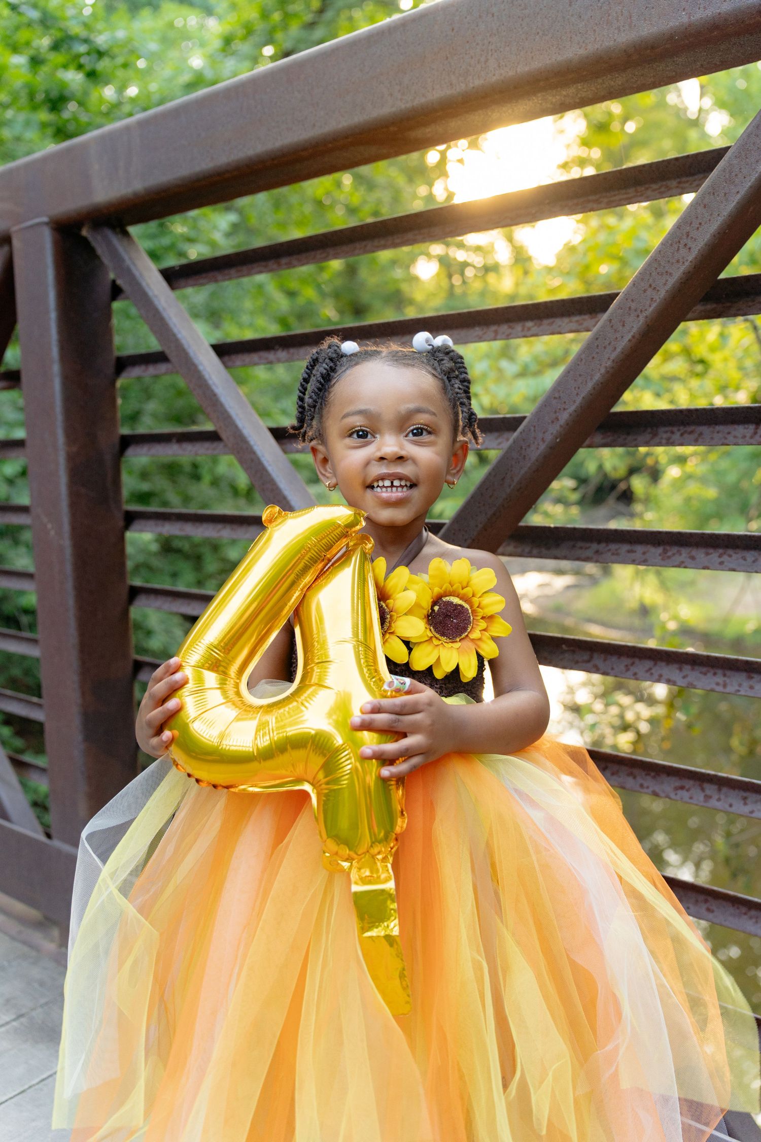 A smiling Black toddler girl in an orange tutu dress holds a gold number 4 balloon and sunflower for her 4th birthday. Taken by Row And Co
