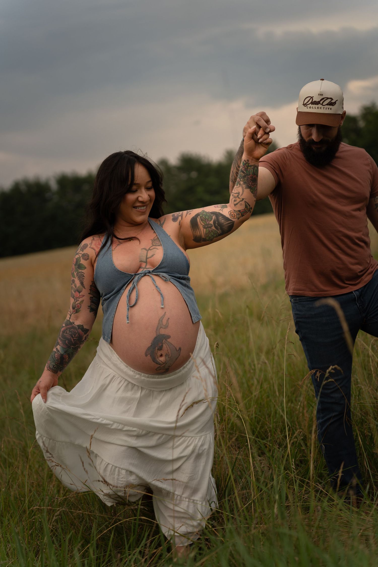 Tattooed pregnant woman in blue crop top and white skirt holds hands with bearded man in field.