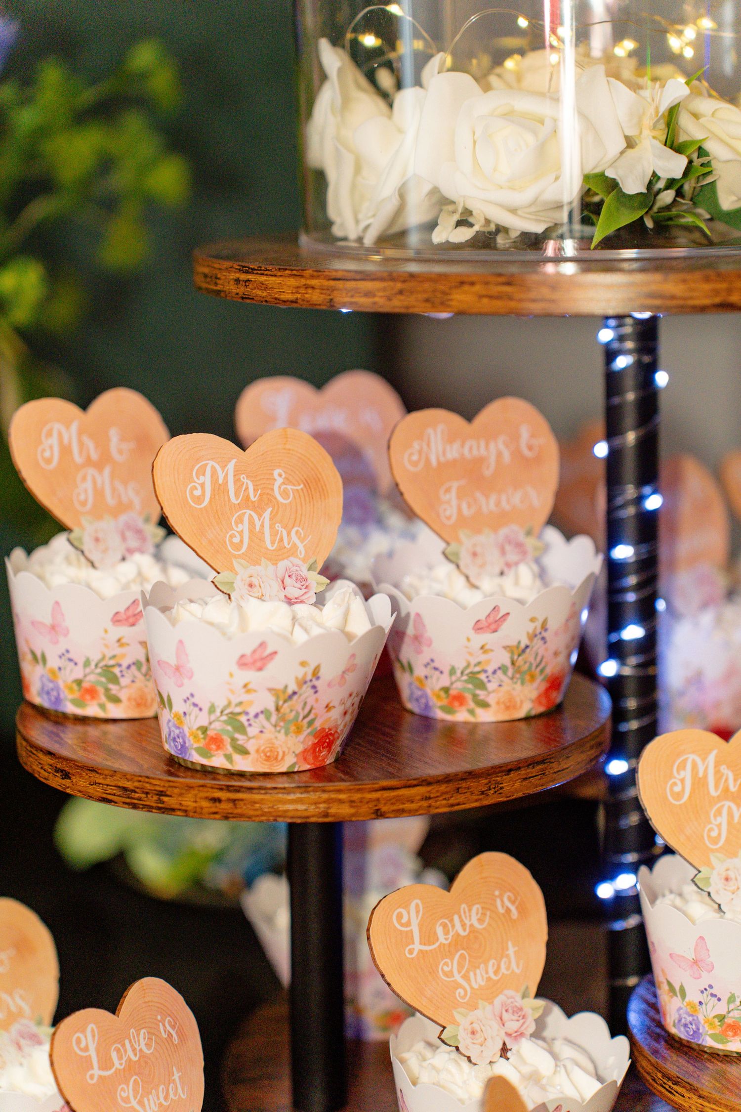Wedding cupcakes with floral wrappers and heart-shaped Mr & Mrs toppers displayed on a rustic wooden tiered stand.