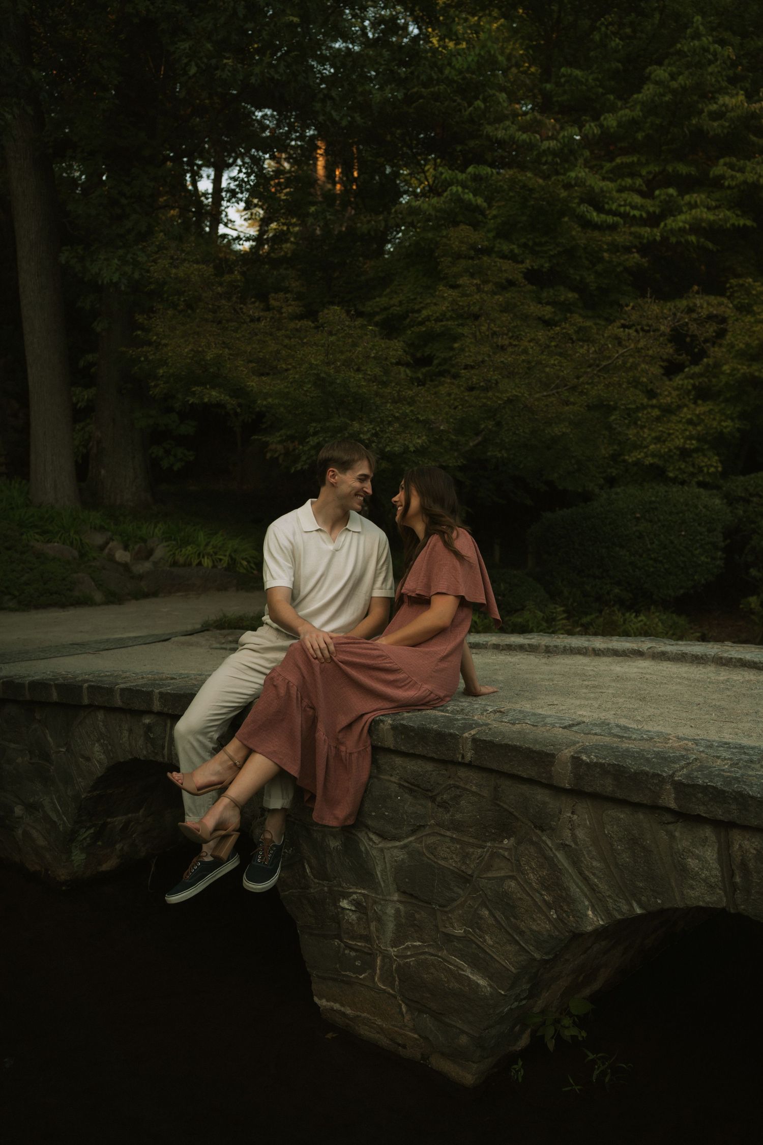Couple sitting closely on a stone bridge at dusk, surrounded by lush green trees, sharing a romantic moment.
