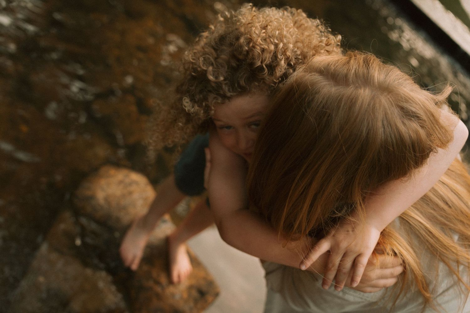 Two young children sharing a tender hug near a rocky stream in a candid outdoor moment. Taken by Row And Co Photography in FXBG VA