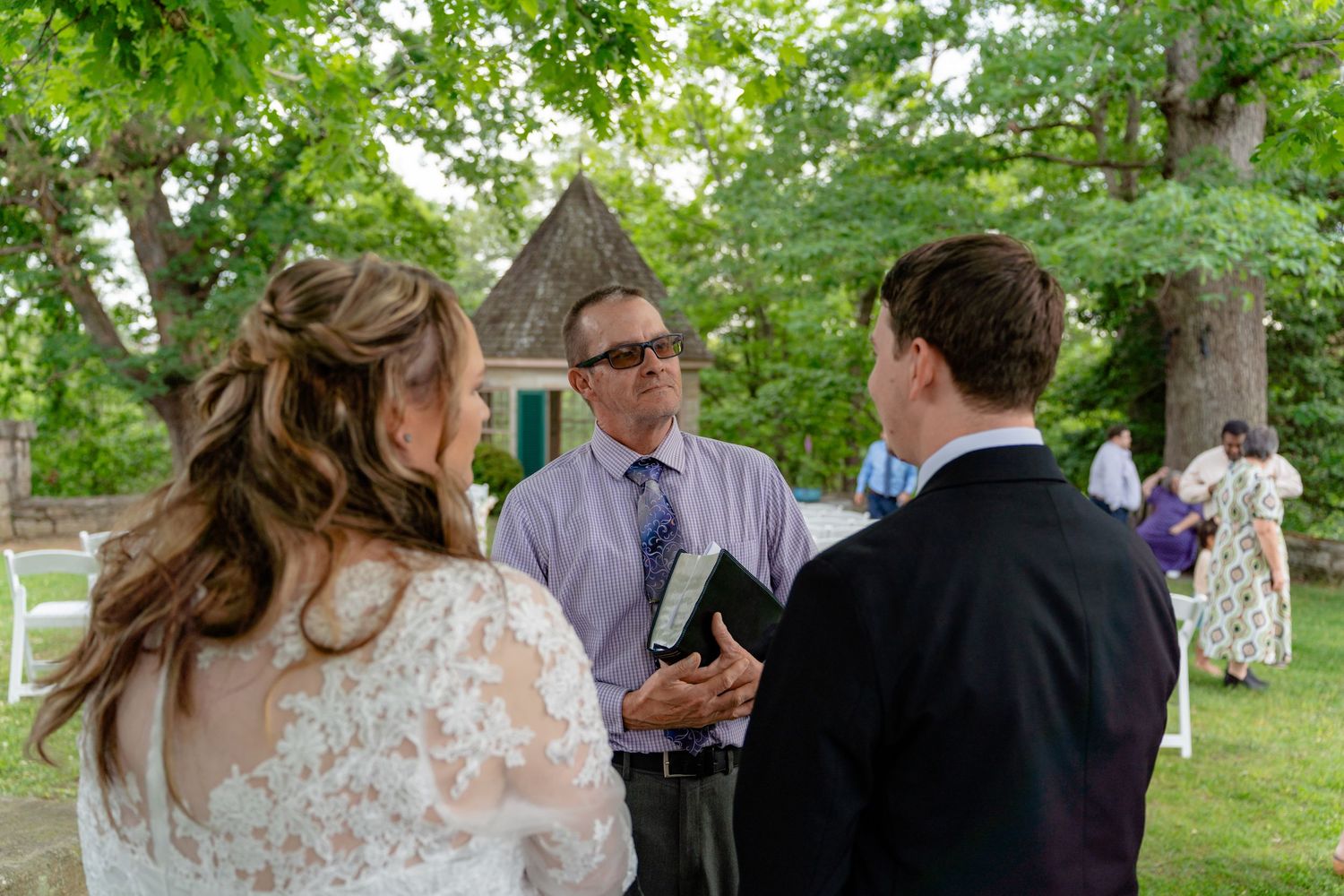 Outdoor wedding ceremony with bride, groom, and officiant holding a book, surrounded by lush green trees.
