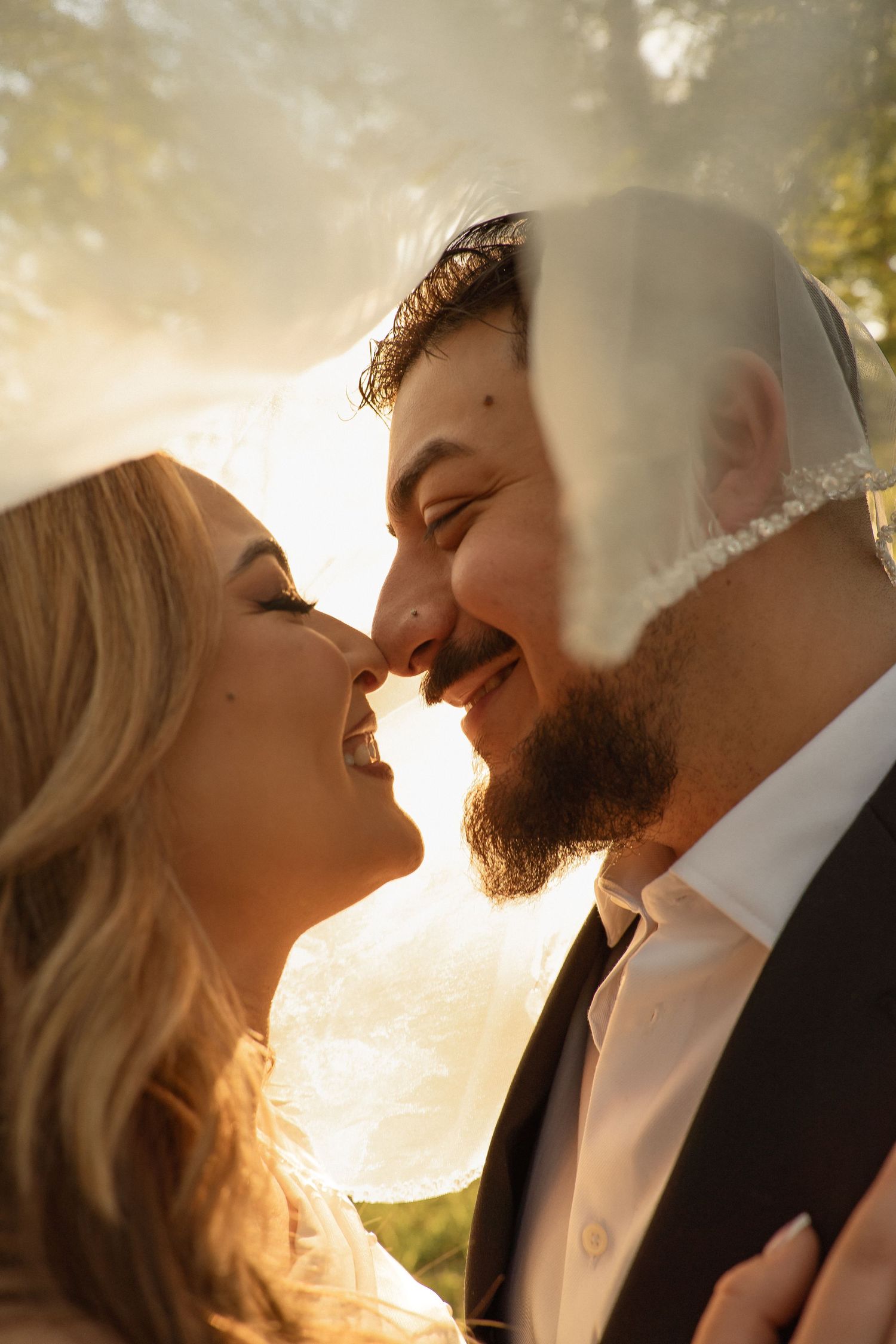 A smiling bride and groom share a romantic nose-to-nose moment under a veil in golden sunset light.
