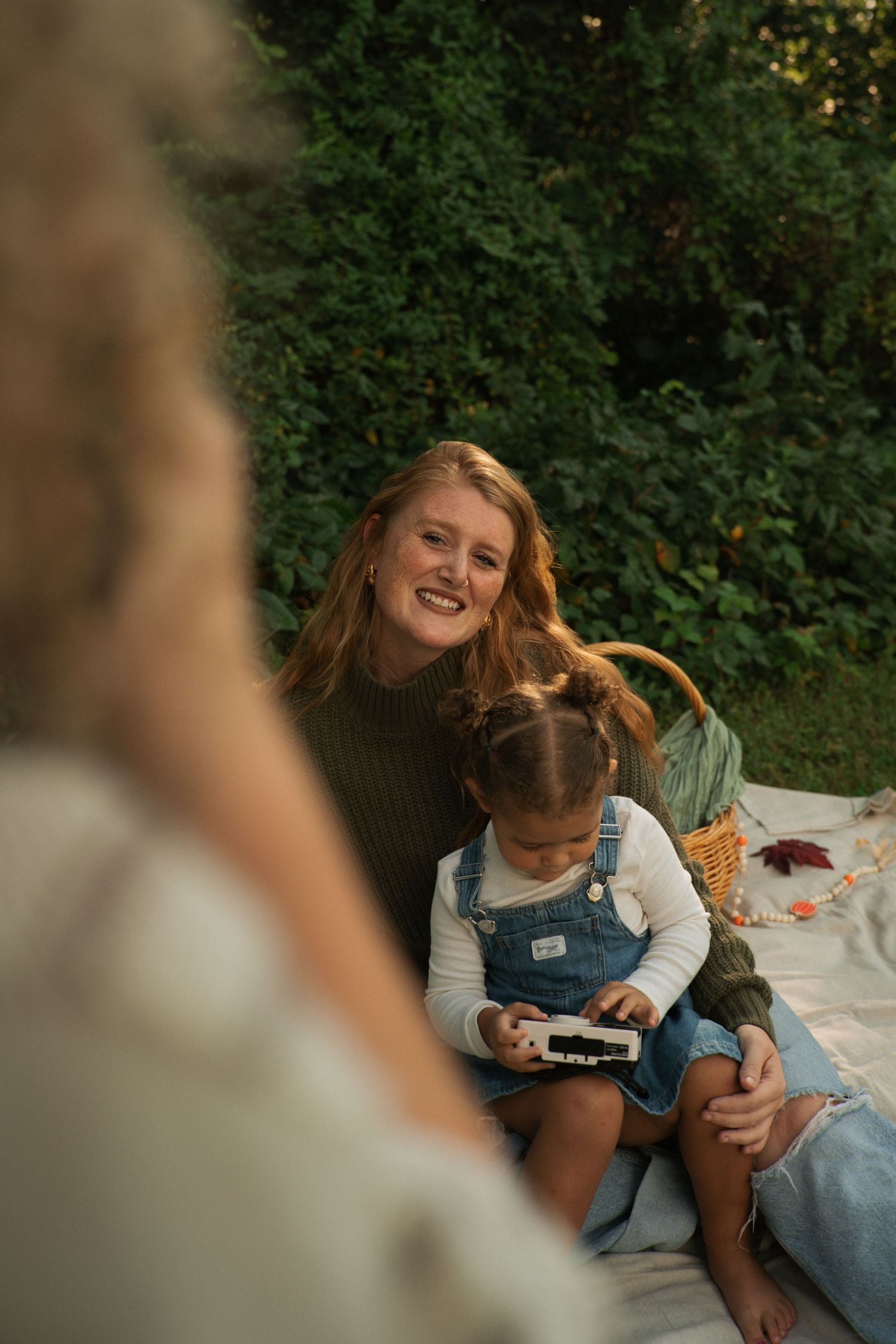 A smiling woman sits on a blanket outdoors with a young girl in denim overalls holding a phone.