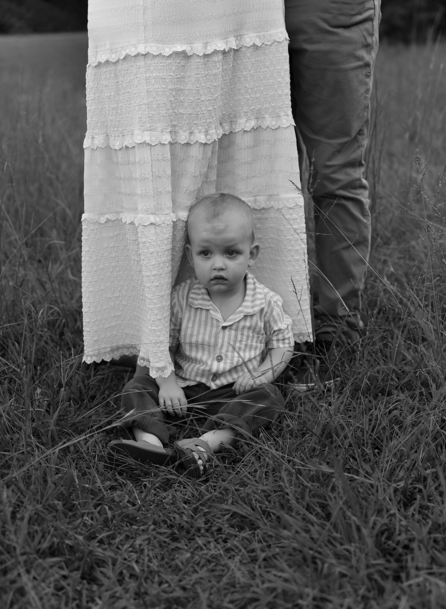 Black and white photo of a baby sitting in grass between parents legs, wearing a striped shirt outdoors.