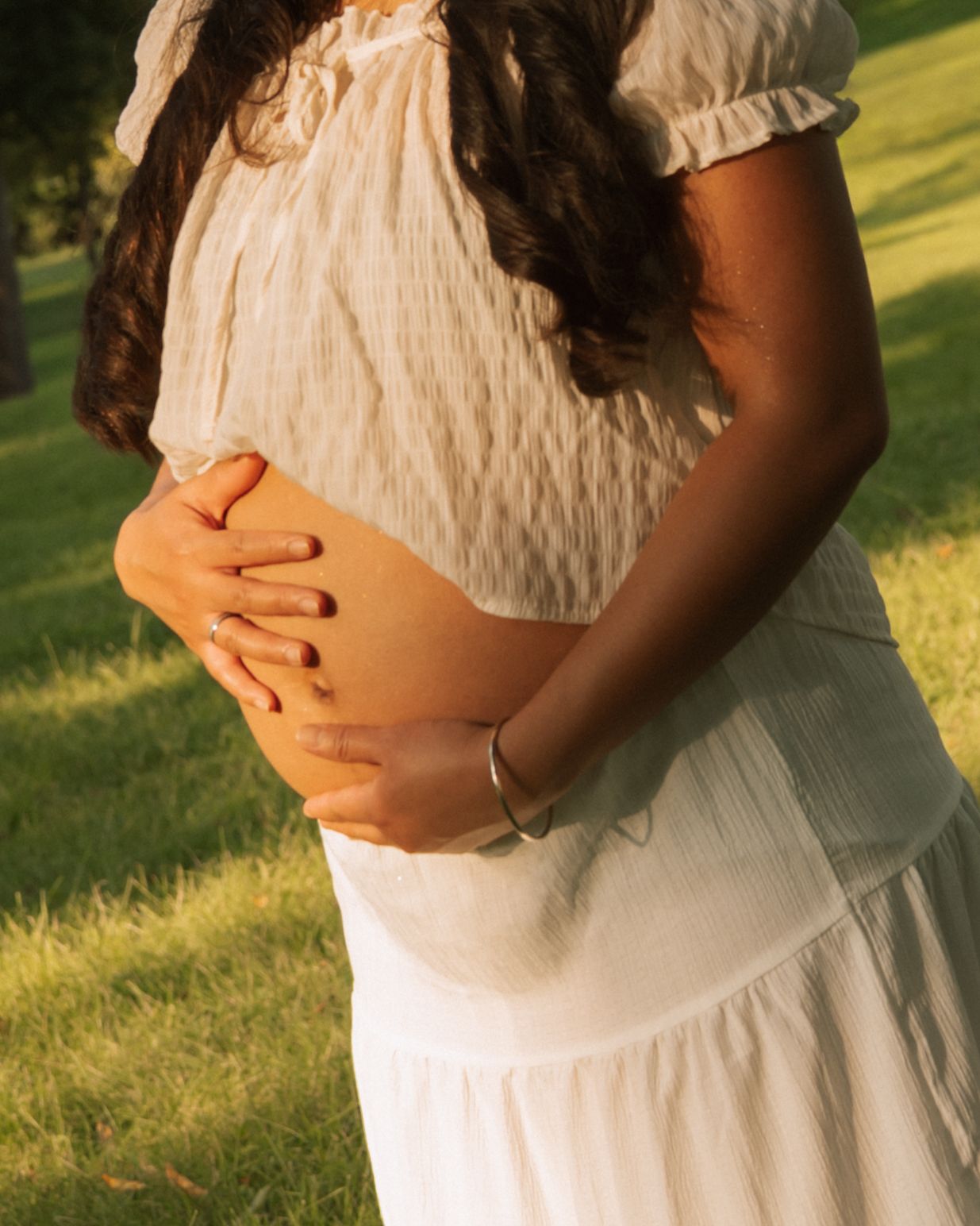 Pregnant woman in white dress cradling baby bump outdoors in sunlit park, wearing silver bracelet.