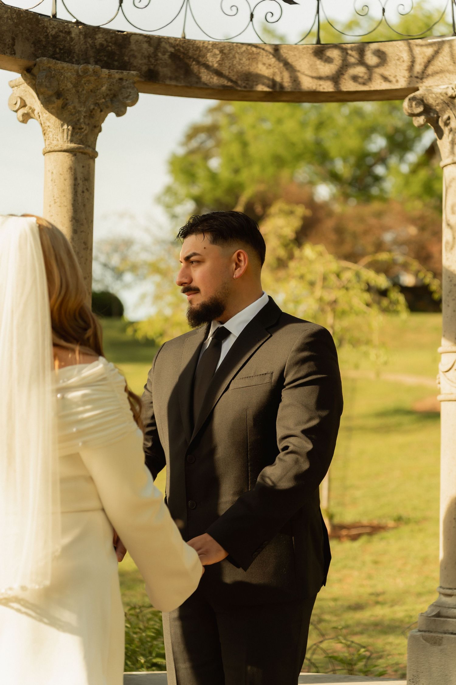 Groom in dark suit holds bride's hands during outdoor wedding ceremony under ornate stone gazebo.