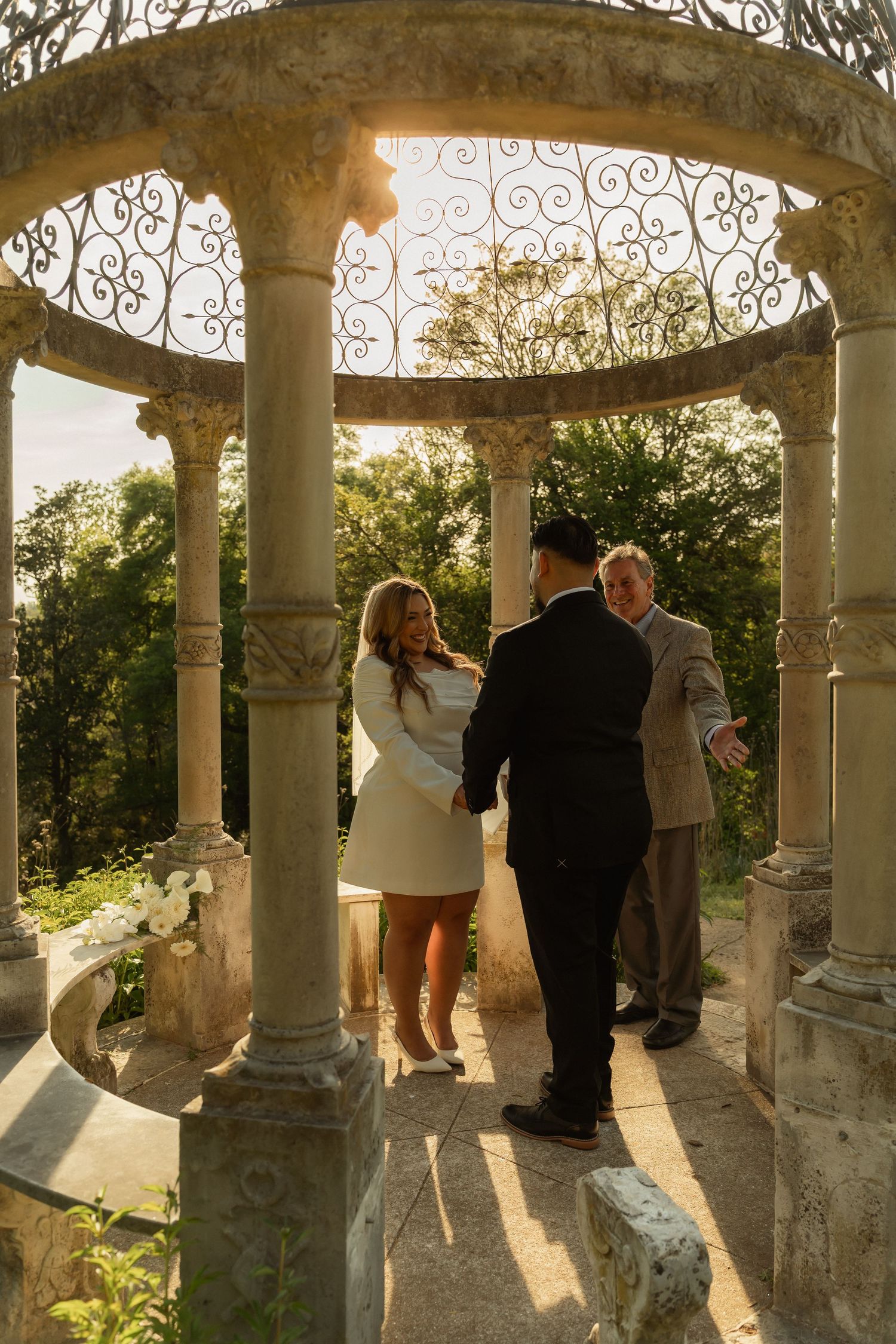 Couple exchanging vows in a sunlit stone gazebo with ornate ironwork, surrounded by lush greenery.