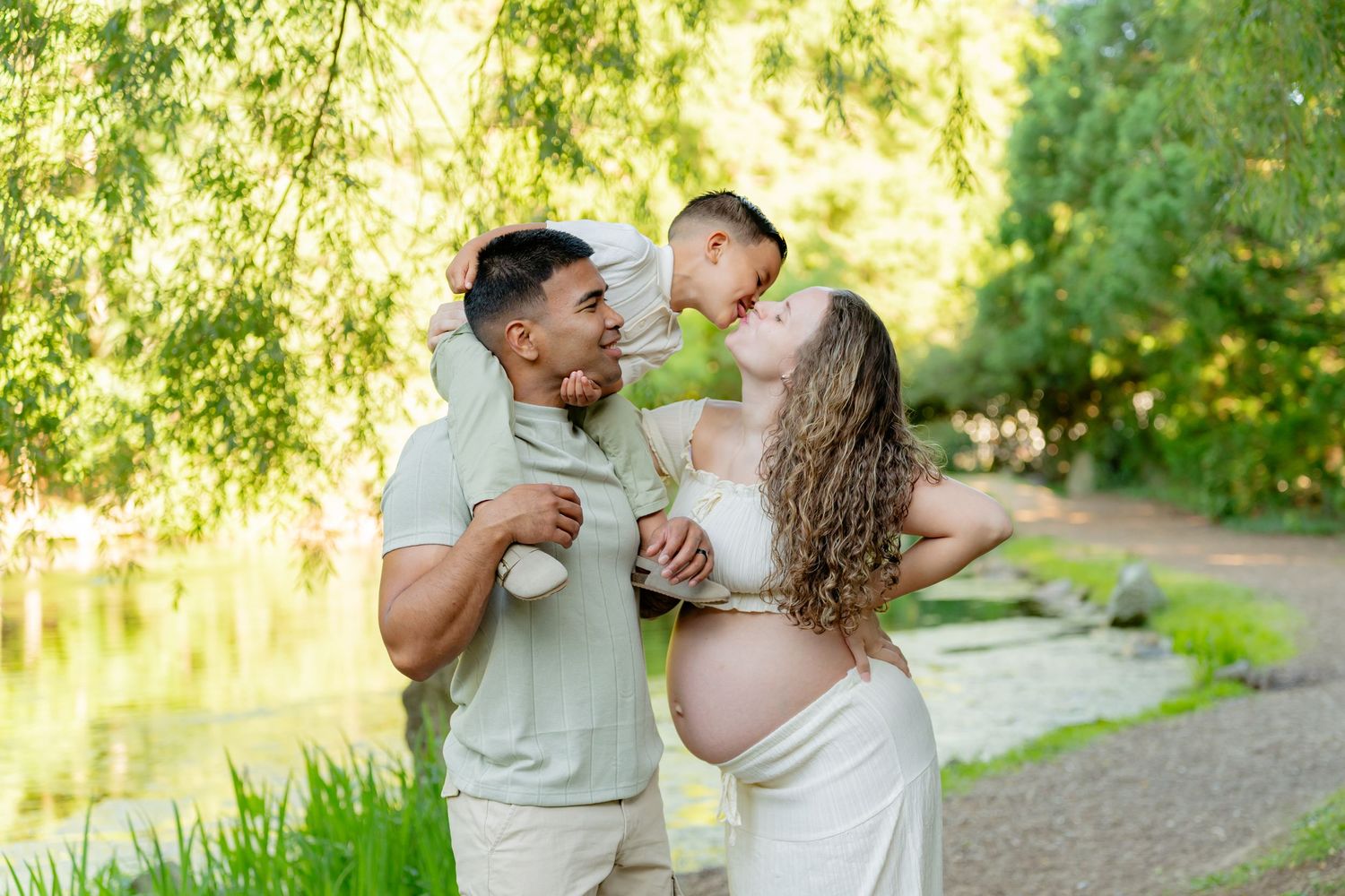 A pregnant couple shares a tender moment with their young son in a lush green park setting. Taken by Row And Co Photography in FXBG VA